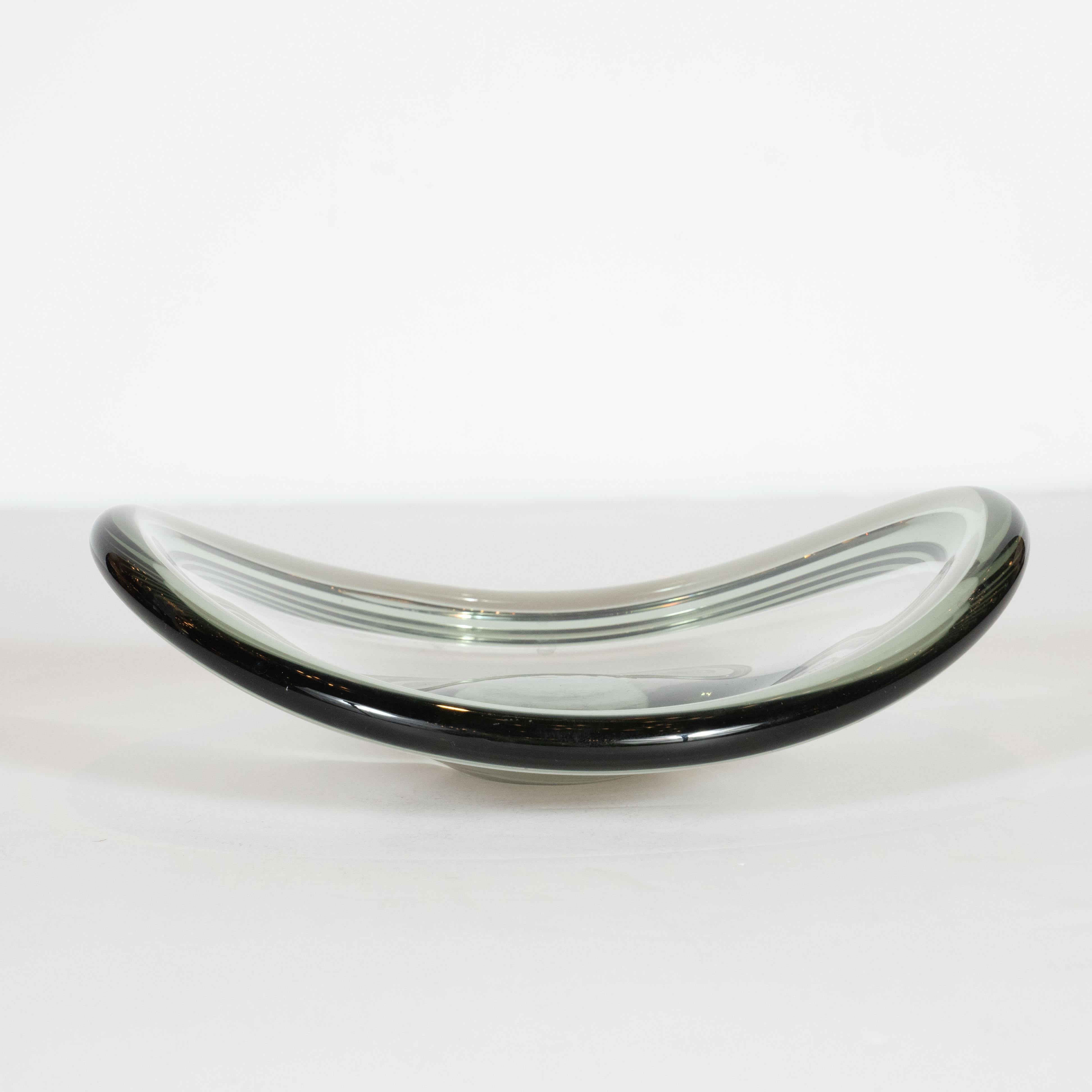 This elegant Mid-Century Modern bowl was hand blown by the celebrated Swedish maker Holmgaard in 1952. It features a sculptural elliptical form with undulating sides and a subtly raised lip all realized in a translucent smoked glass. With its clean