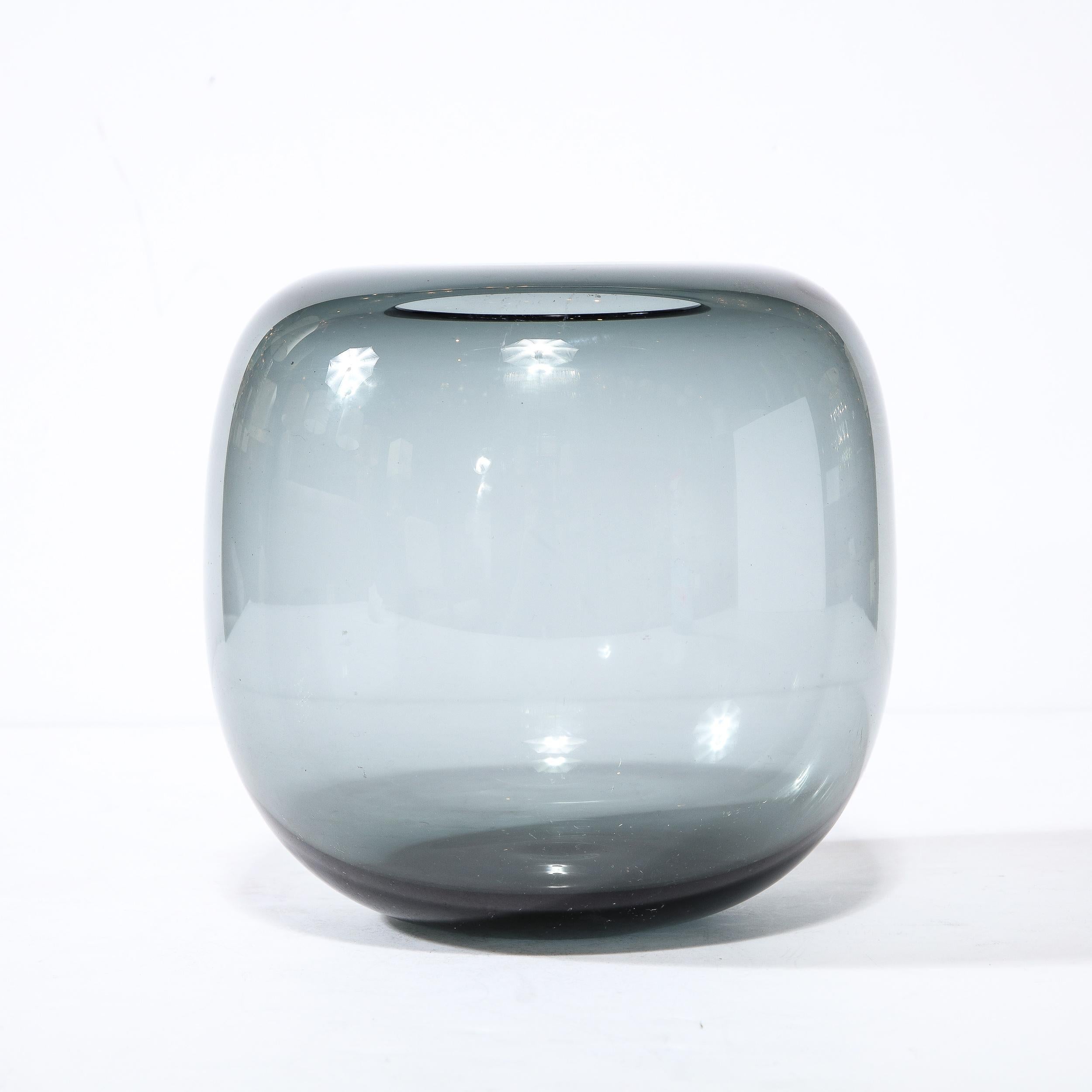 This elegant Mid Century Modern vase was realized by the esteemed maker Holmegaard in Denmark circa 1960. It offers an elongated spherical form with a rounded shoulders and a circular mouth- all in a subtle smoked translucent glass. This piece is a