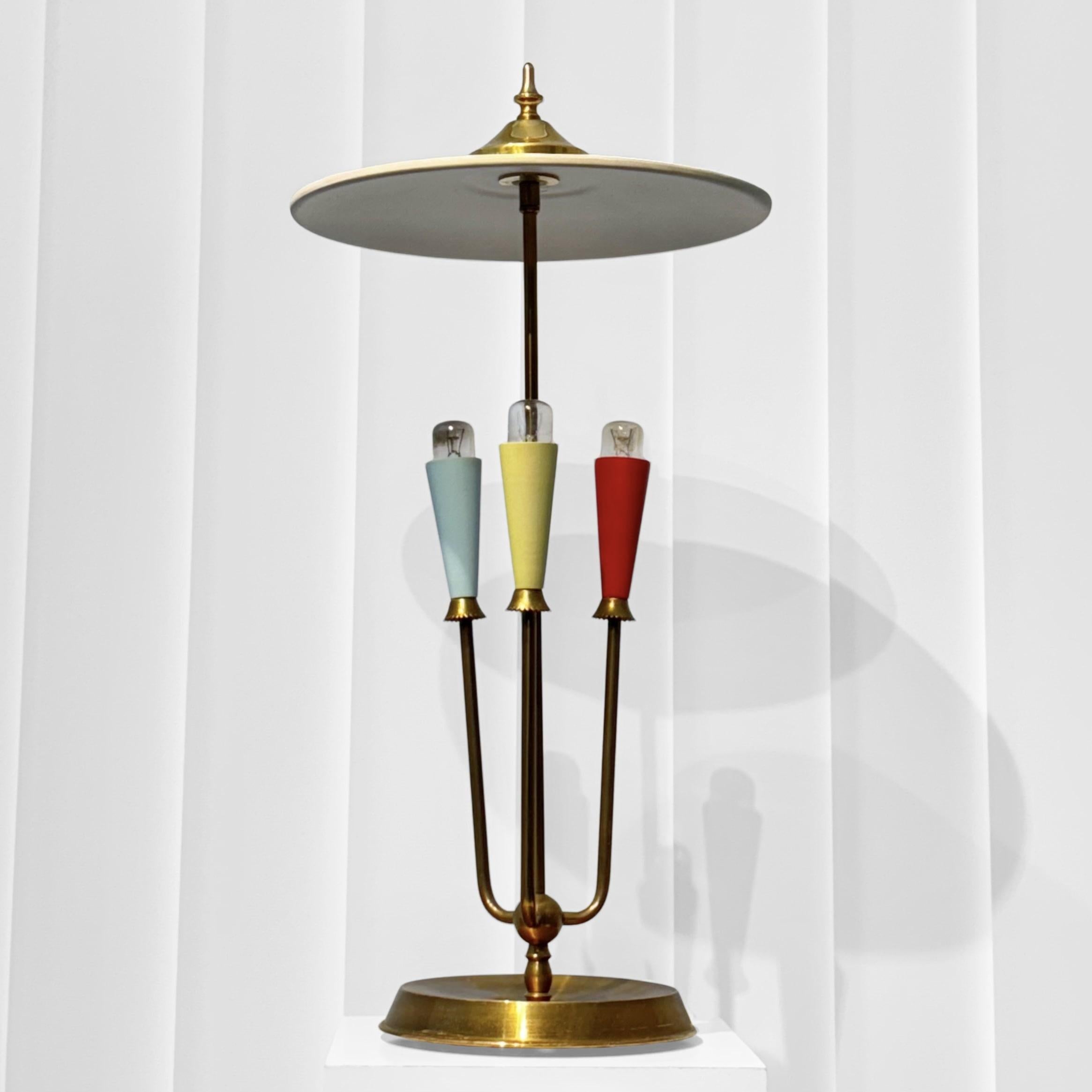 Lampe de table moderniste du milieu du siècle, fabriquée en laiton, avec trois bras sur une base circulaire. Chaque bras porte un porte-lampe conique de couleur bleue, verte et rouge, qui confère à la lampe son caractère distinctif. Au sommet, un