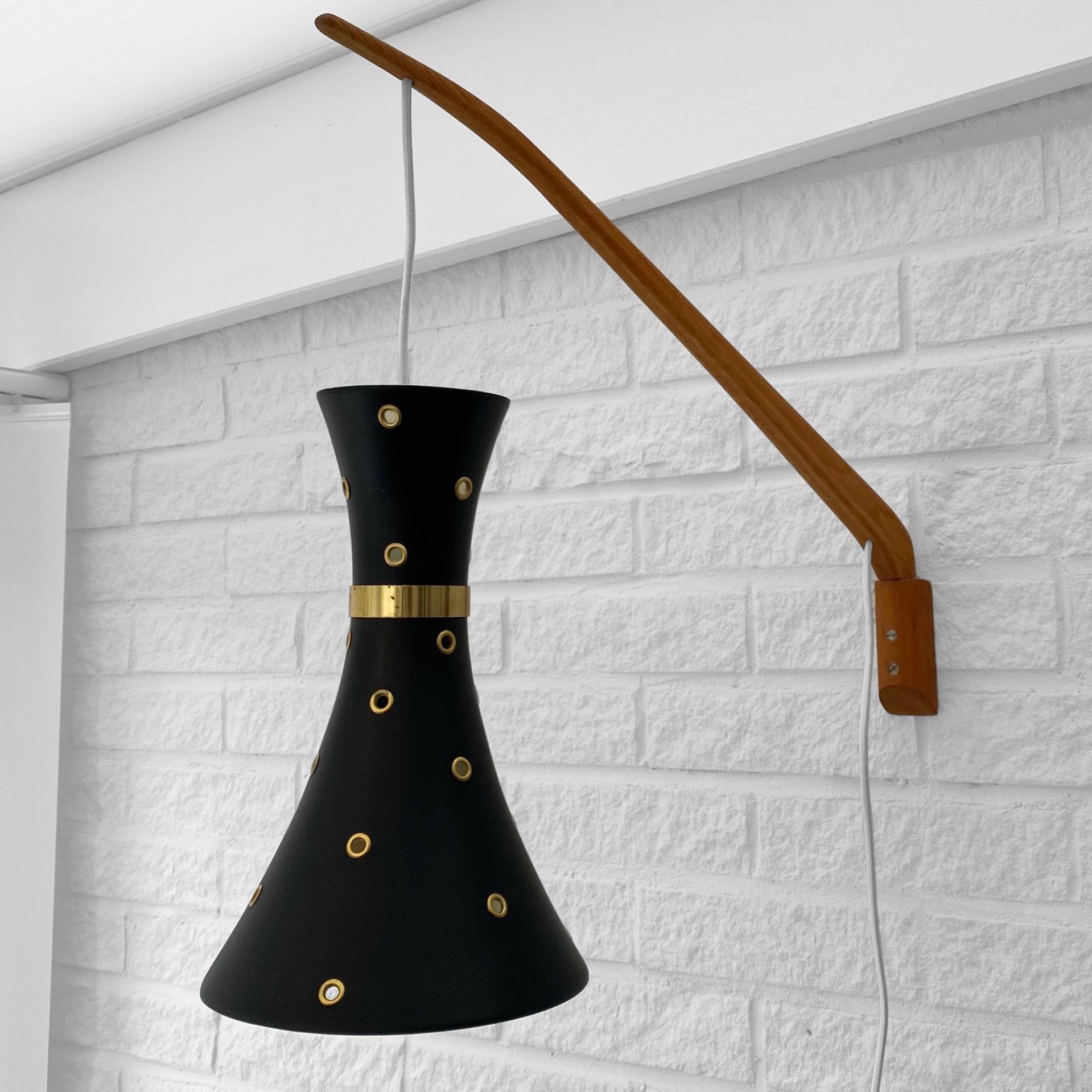 A Scandinavian mid-century wall lamp, crafted from oak and metal with a slender wooden arm. Black hourglass-shaped metal shade with perforated dots and brass details. Produced by an unidentified Swedish manufacturer in the 1950s. Length of the arm