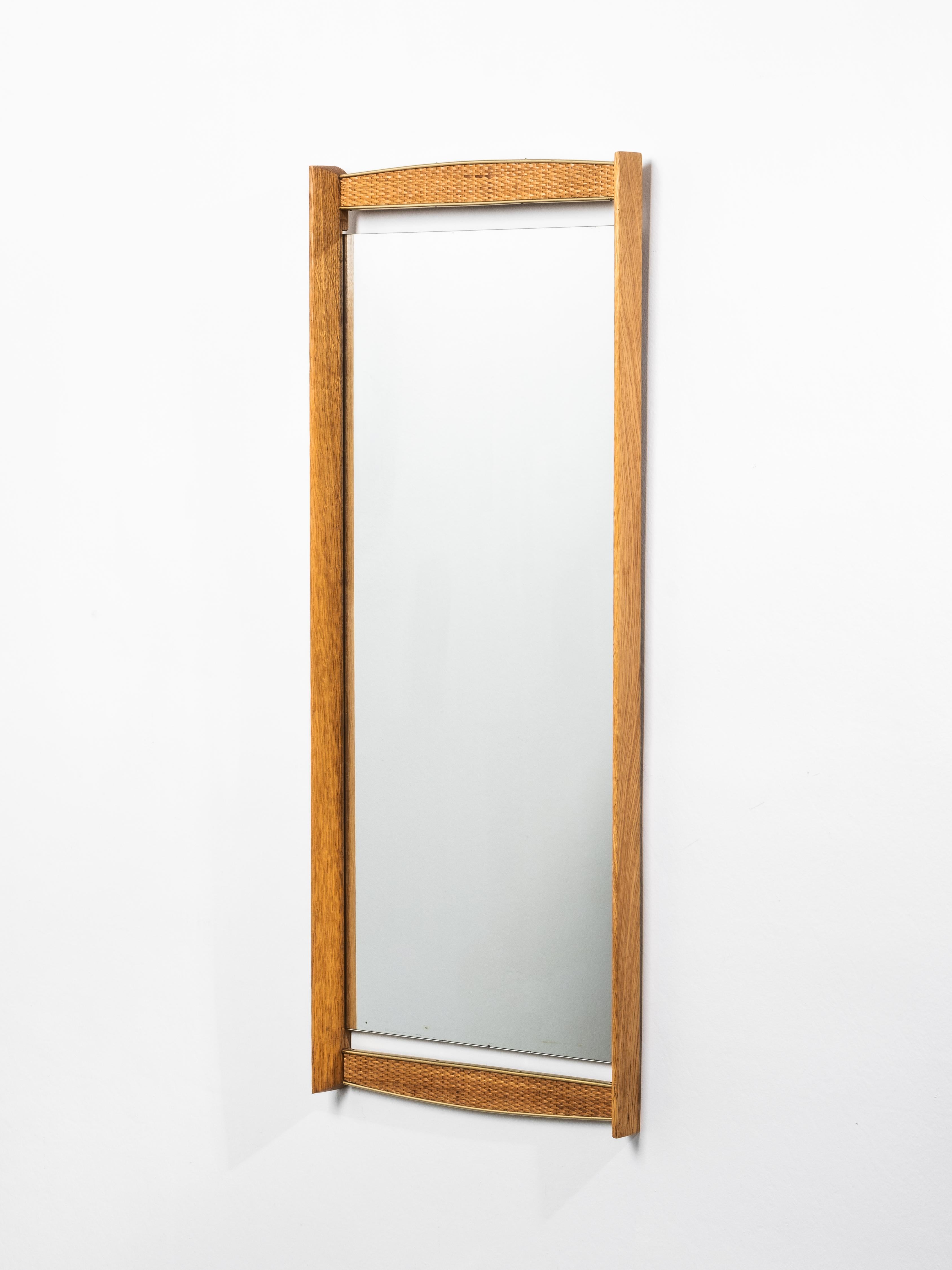 Miroir mural scandinave raffiné du milieu du siècle, fabriqué en chêne massif avec d'élégants panneaux en rotin tressé encadrés de laiton. Le design présente des bords supérieur et inférieur subtilement arqués, créant une silhouette gracieuse et