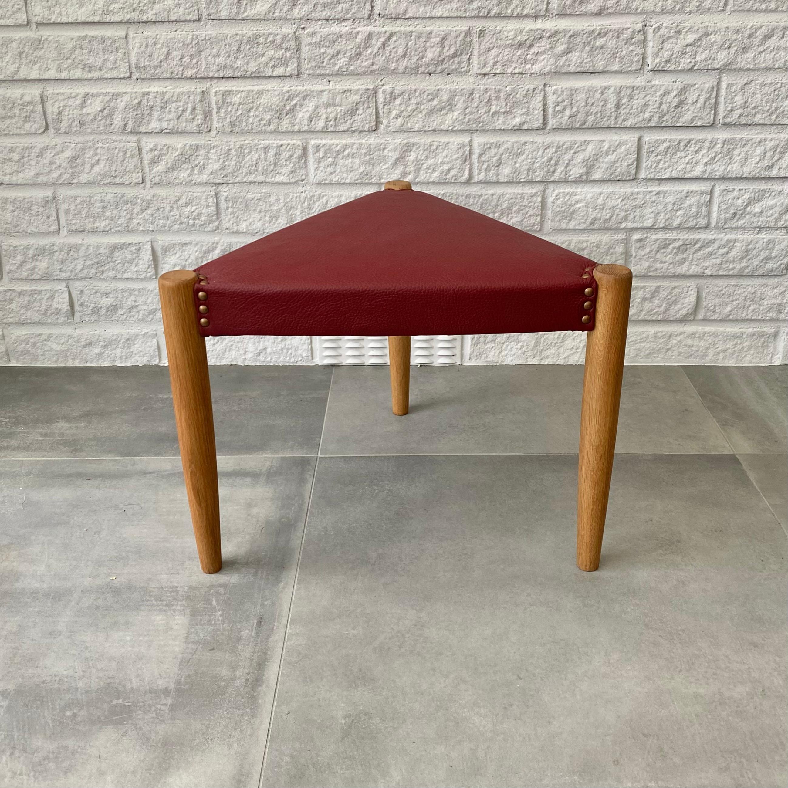 Scandinavian mid-century stool made from solid oak with a triangular seat upholstered in red leather. Decorative brass rivets surrounding the top of each of the three leg. Designed and produced by an unidentified Swedish manufacturer in the 1960s.
