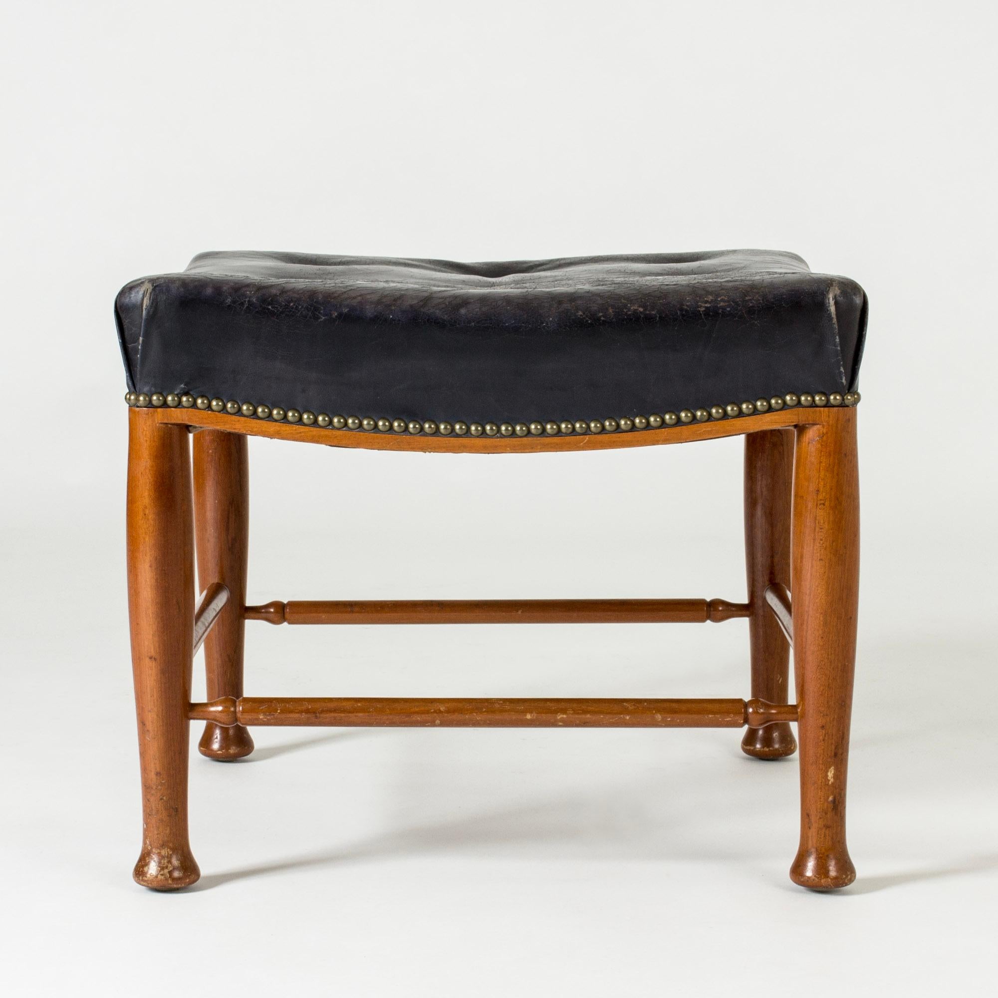 Ottoman by Josef Frank, made from mahogany with elegantly sculpted legs. Original black leather upholstery, decorative brass nails around the edge of the seat.