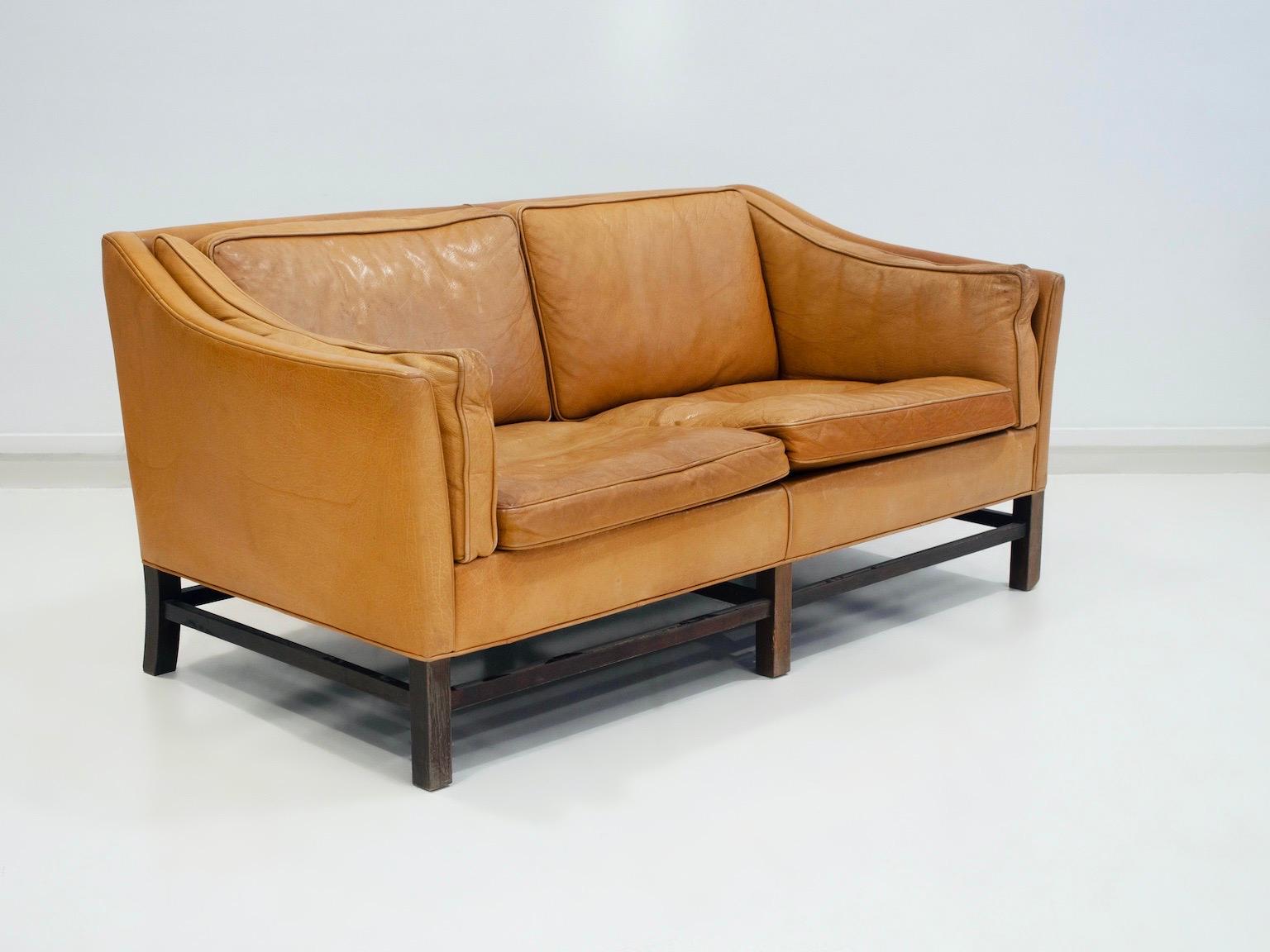 Two-seater caramel brown leather sofa designed in Denmark and manufactured by Grant in circa 1970/80s. Mahogany wood base and leather upholstery with patina due to age and use. Seat and backrest with loose cushions. Comfortable sofa that combines