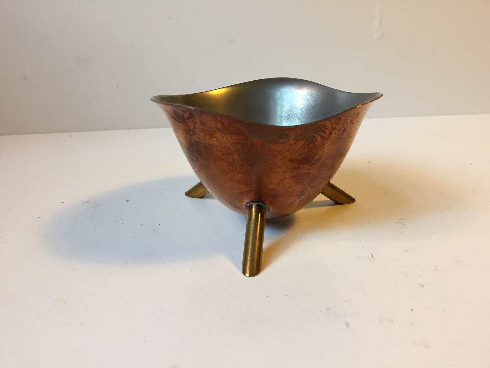 A small organically shaped nut or candy bowl fashioned out of a bended copper plate that has been linned with pewter. The 'legs' are made from brass. It was designed anonymously in Scandinavia during the 1950s in a style reminiscent of Henning