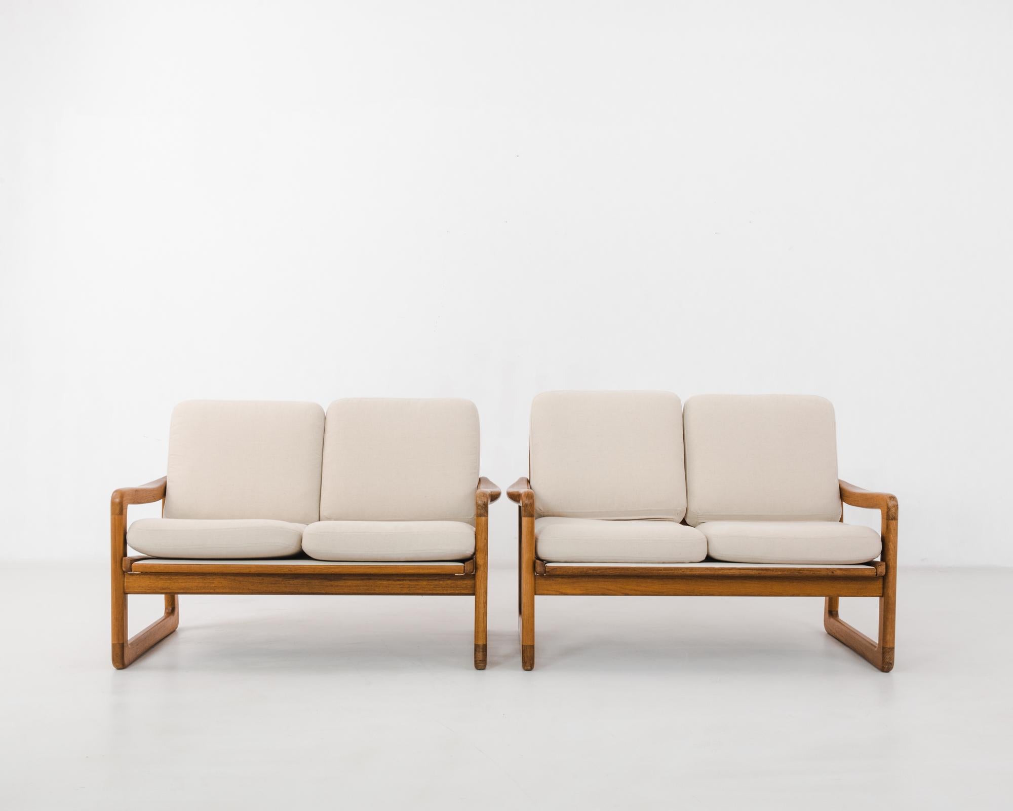 A pair of teak sofas from 1970s Denmark. The clean lines of the frame evoke a sense of stylish ease, characteristic of Scandinavian modernist design. Seat cushions are upholstered in a natural white, creating a fresh contrast with the warm polish of
