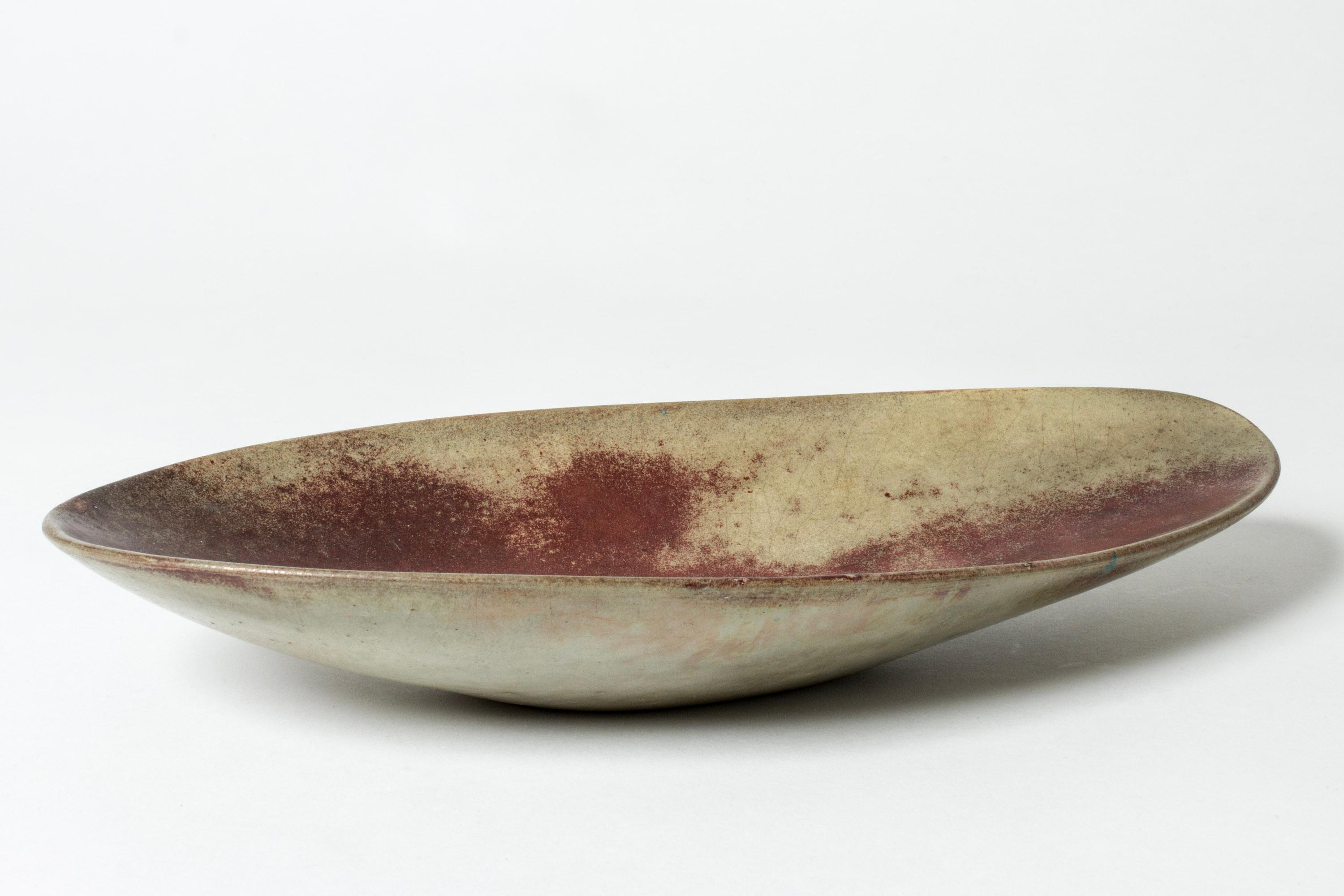 Platter by Hans Hedberg, made in faience. Smooth, organic form with striking oxblood glaze on a grey background.