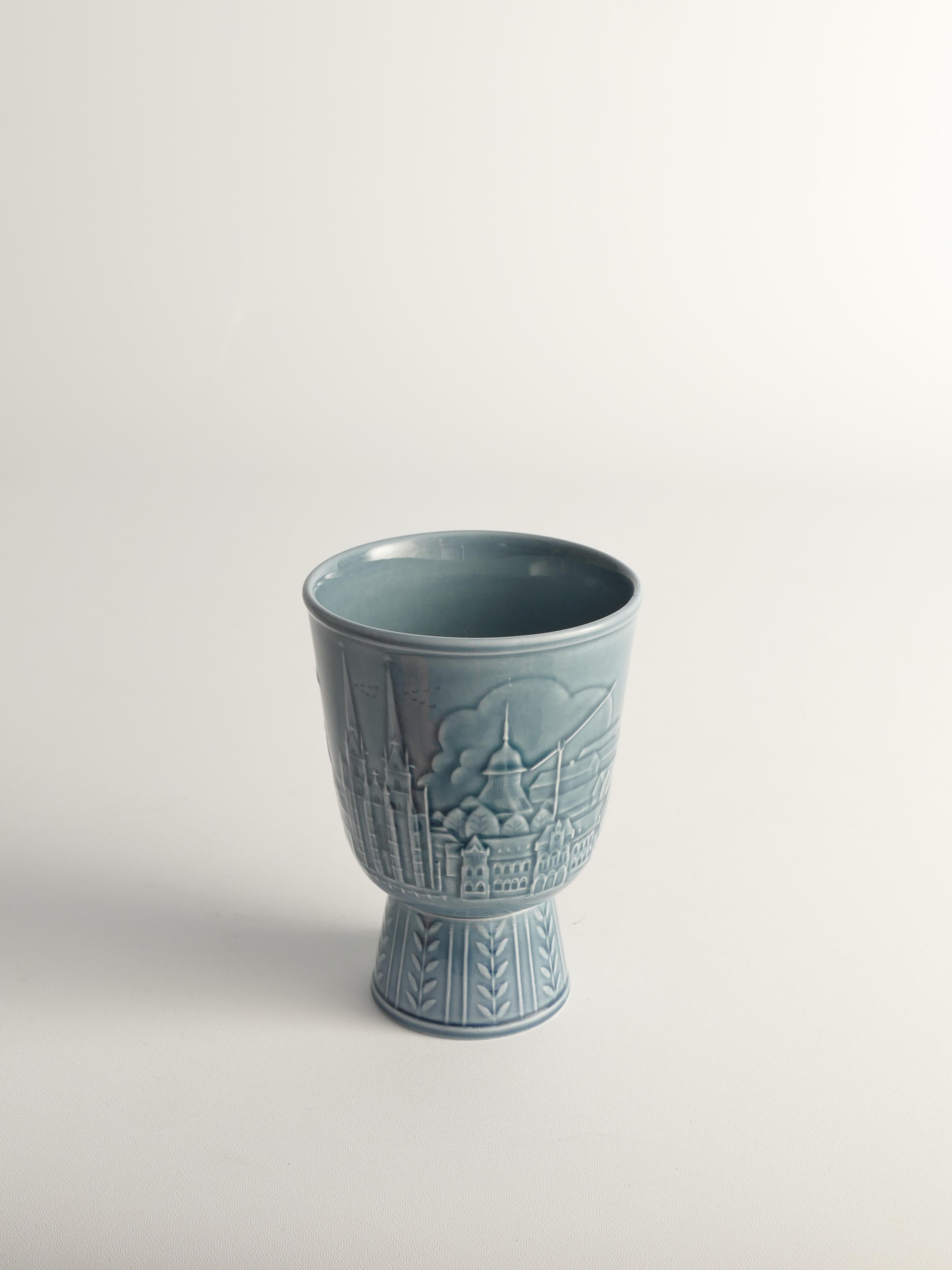 Questo vaso moderno scandinavo, disegnato da Gunnar Nylund per Rörstrand, raffigura l'affascinante cittadina di Skara con la sua iconica chiesa, gli uccelli che si librano nel cielo e il pittoresco paesaggio di case, alberi, campi e cespugli. Il