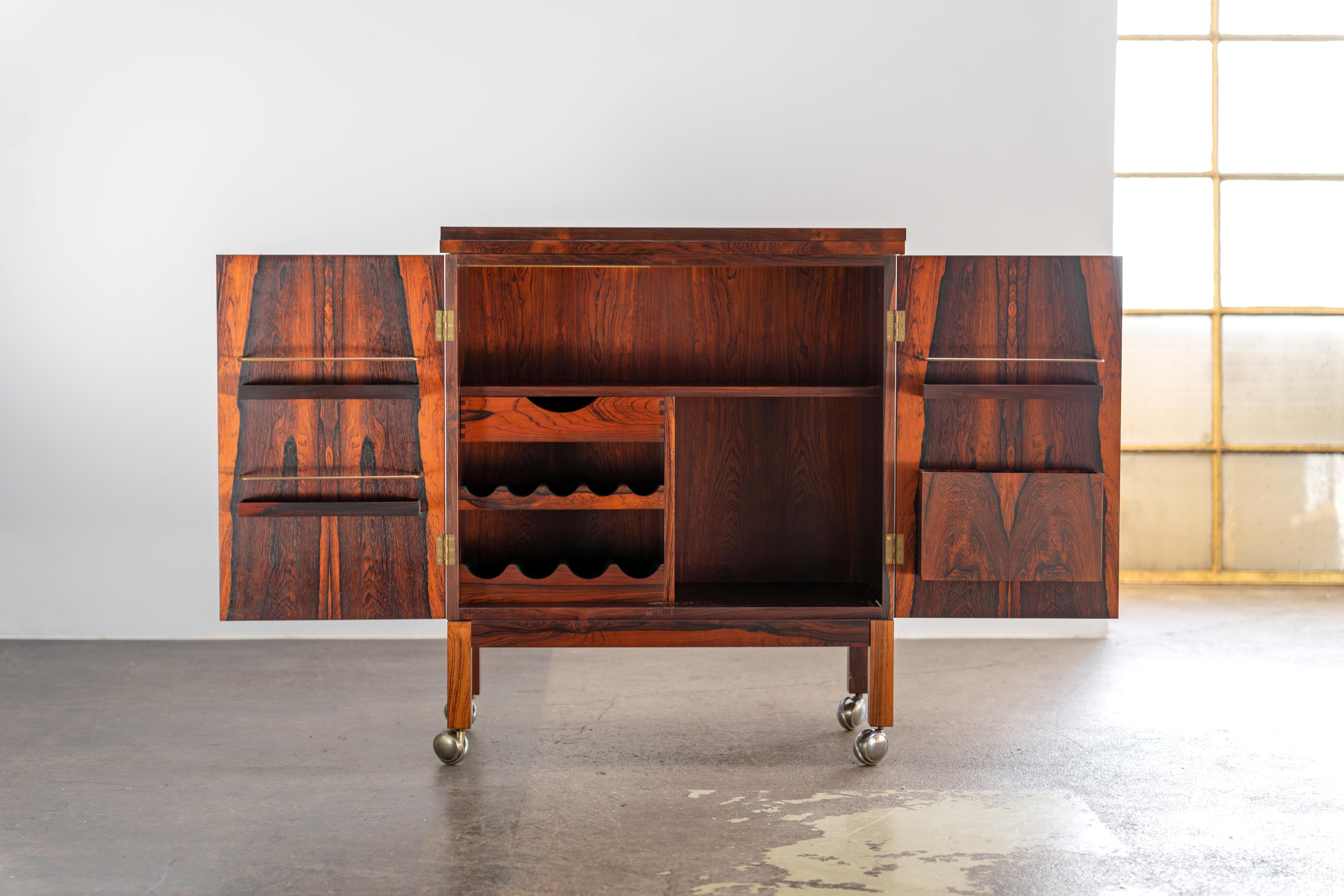 Disegnato per Bruksbo, questo raro mobile bar in legno di palissandro colpisce per il suo piano pieghevole, le cerniere in ottone e gli interni ben studiati con ripiani, un portabottiglie rivestito di verde e portabicchieri. Le rotelle cromate