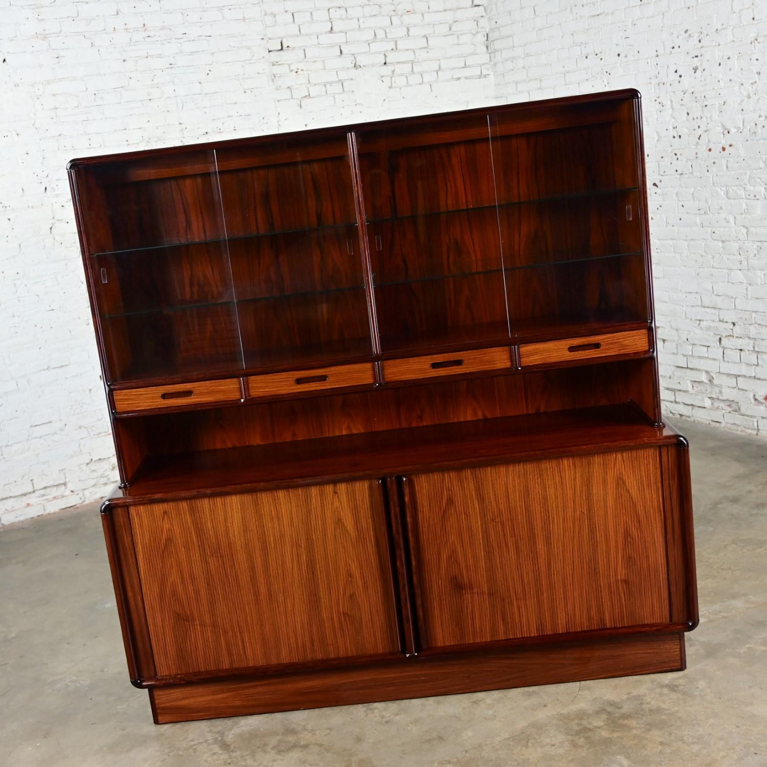 Handsome vintage Scandinavian Modern Rosewood lighted China hutch or sideboard by Kibaek Mobelfabrik A/S Made in Denmark. Comprised of 2 pieces including a top hutch with glass shelves & glass bypass doors and a lower buffet with tambour doors, wood