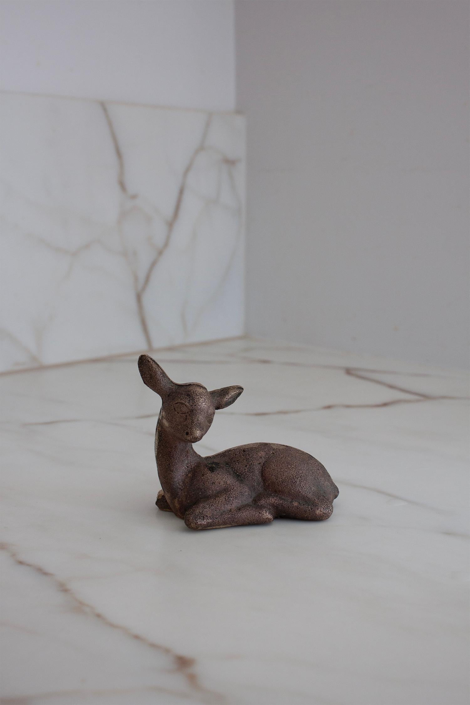 La escultura muestra una figura yacente de cervatillo, hecha de latón fundido patinado con textura de bronce. La figura tiene una forma simplificada, casi modernista, con un rostro ligeramente abstracto y líneas suaves, que era el estilo típico de