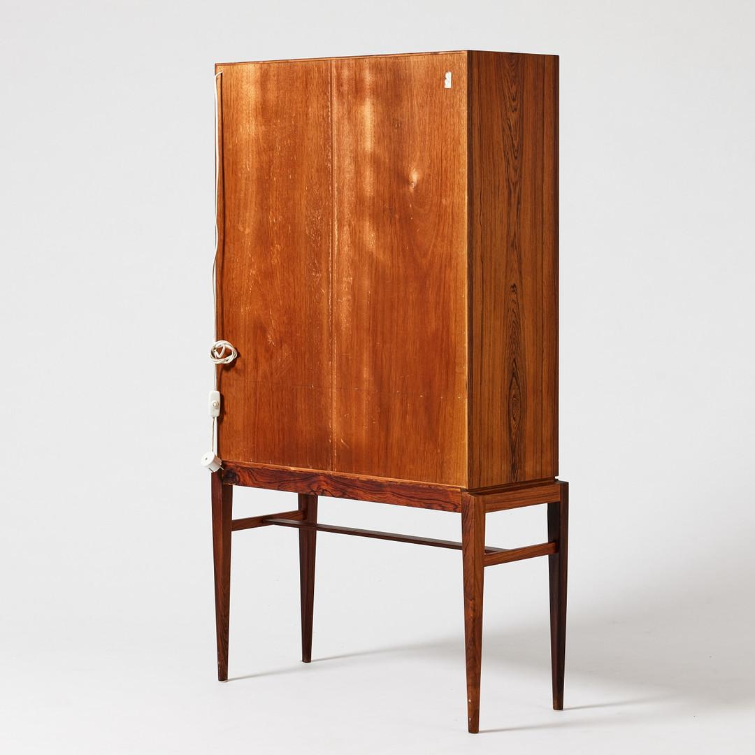 Descrizione
Impiallacciato in legno duro, di forma rettangolare su supporto a gambe accoppiate, ha cinque cassetti, ante scorrevoli in vetro, interno con due ripiani e illuminazione.
Altezza 140,5 cm, L78 x profondità 31 cm

LETTERATURA 
Andreas