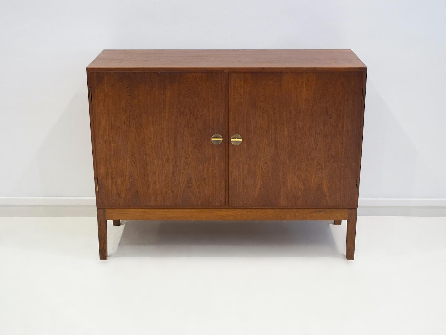 Cabinet of teak, later mounted on unoriginal oak feet of slightly lighter color. Front with two doors. Interior with two shelves and four pullout trays. Manufactured in Denmark by FDB Møbler. Two keys that serve as handles included.