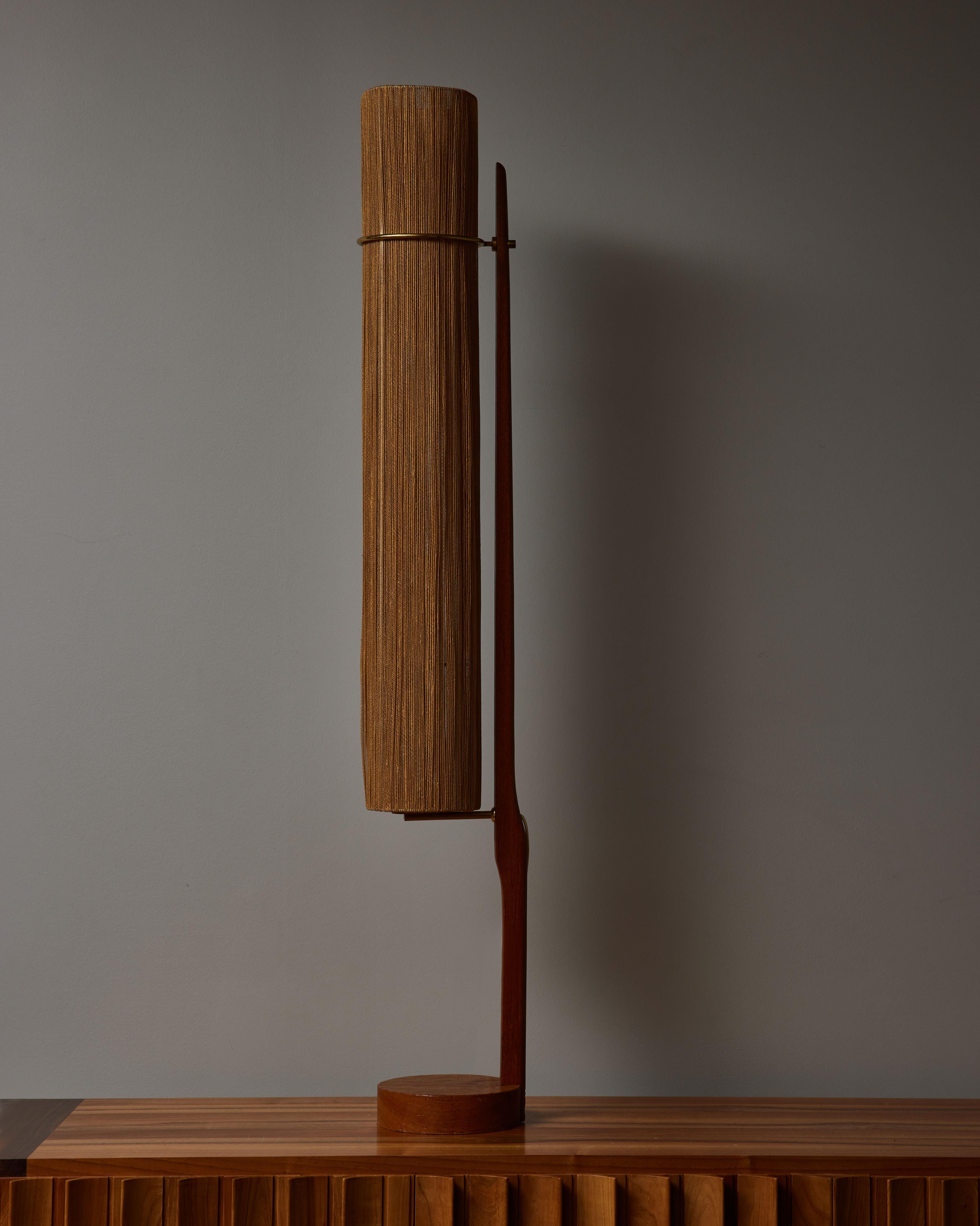 Lampadaire en teck composé d'un pied circulaire, d'une tige verticale décalée avec un anneau en laiton poli, supportant un abat-jour étroit en cordes de coton.
