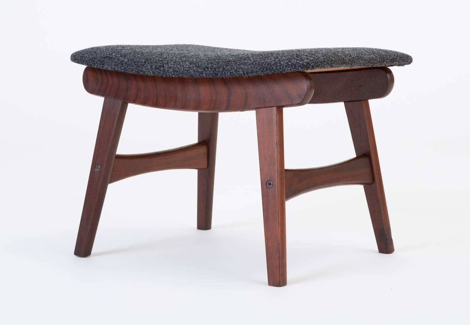 A modest Scandinavian ottoman or footrest has a base of teak wood with flat, angled legs and sculpted cross-supports. The curved cushion is upholstered in a heathered midnight blue textile that sits on slightly bowed teak runners. Unsigned.