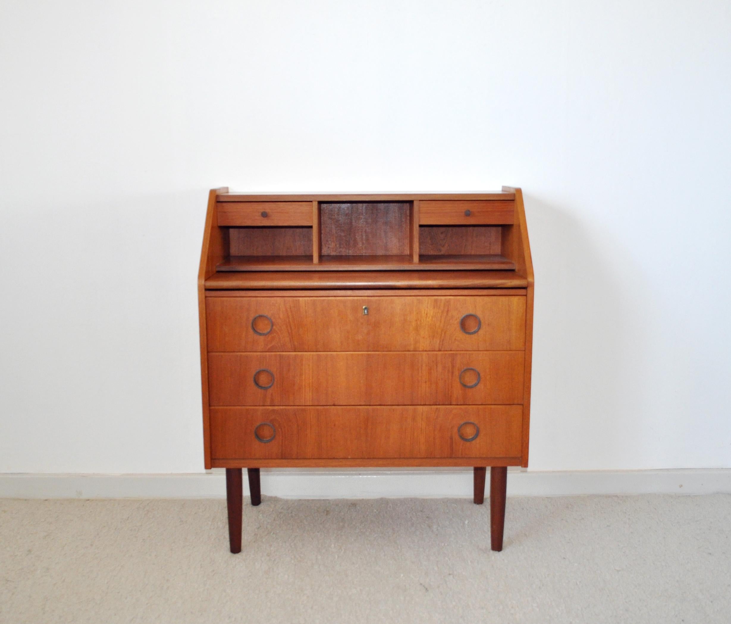 Swedish Secretaire or chest of drawers, 1970s.
Teak veneer and legs made of solid teak.
The top has cubby holes and 2 small drawers. Lower section has three large drawers with round plastic handles. One divided and painted. The desk can be pulled