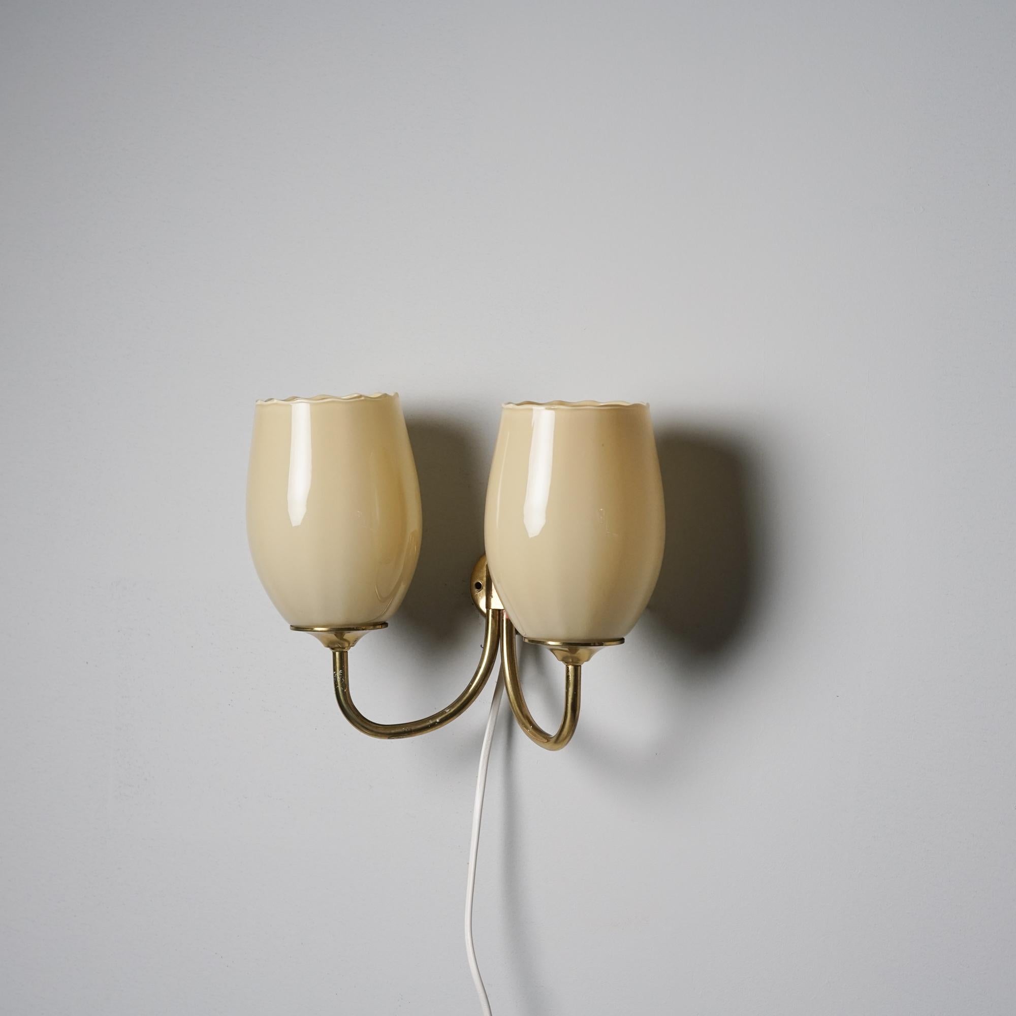 Lampada da parete a due bracci, prodotta in Finlandia, anni '50. Struttura in ottone, paralumi in vetro opalino. Buone condizioni vintage, lieve patina coerente con l'età e l'uso. 