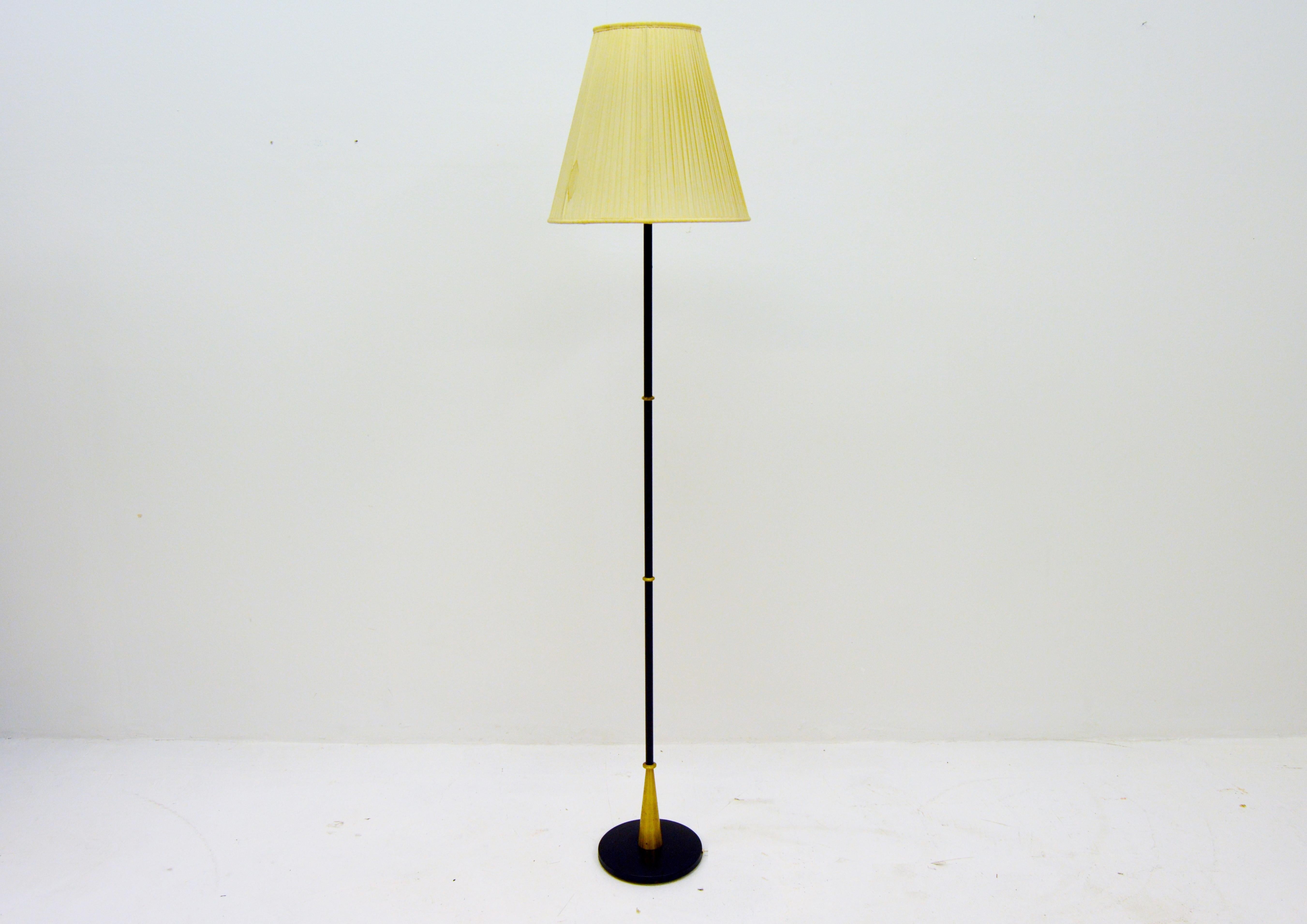 Unusual floor lamp with black metal bar and brass details. The brass at the foot forms a small cone and the rings make the lamp distinguish from other lamps from the same period of time. The top is an uplight made of glass and the lampshade is