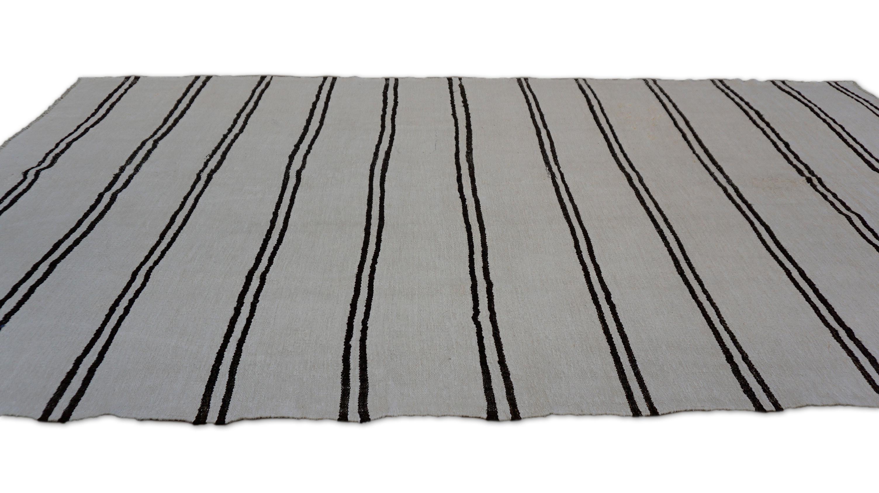 Questo kilim di canapa vintage tessuto a mano presenta un'estetica audace e senza tempo grazie alle sue sorprendenti strisce orizzontali nere su un fondo di canapa naturale e non tinta. Il tappeto incarna il minimalismo con carattere: il suo motivo