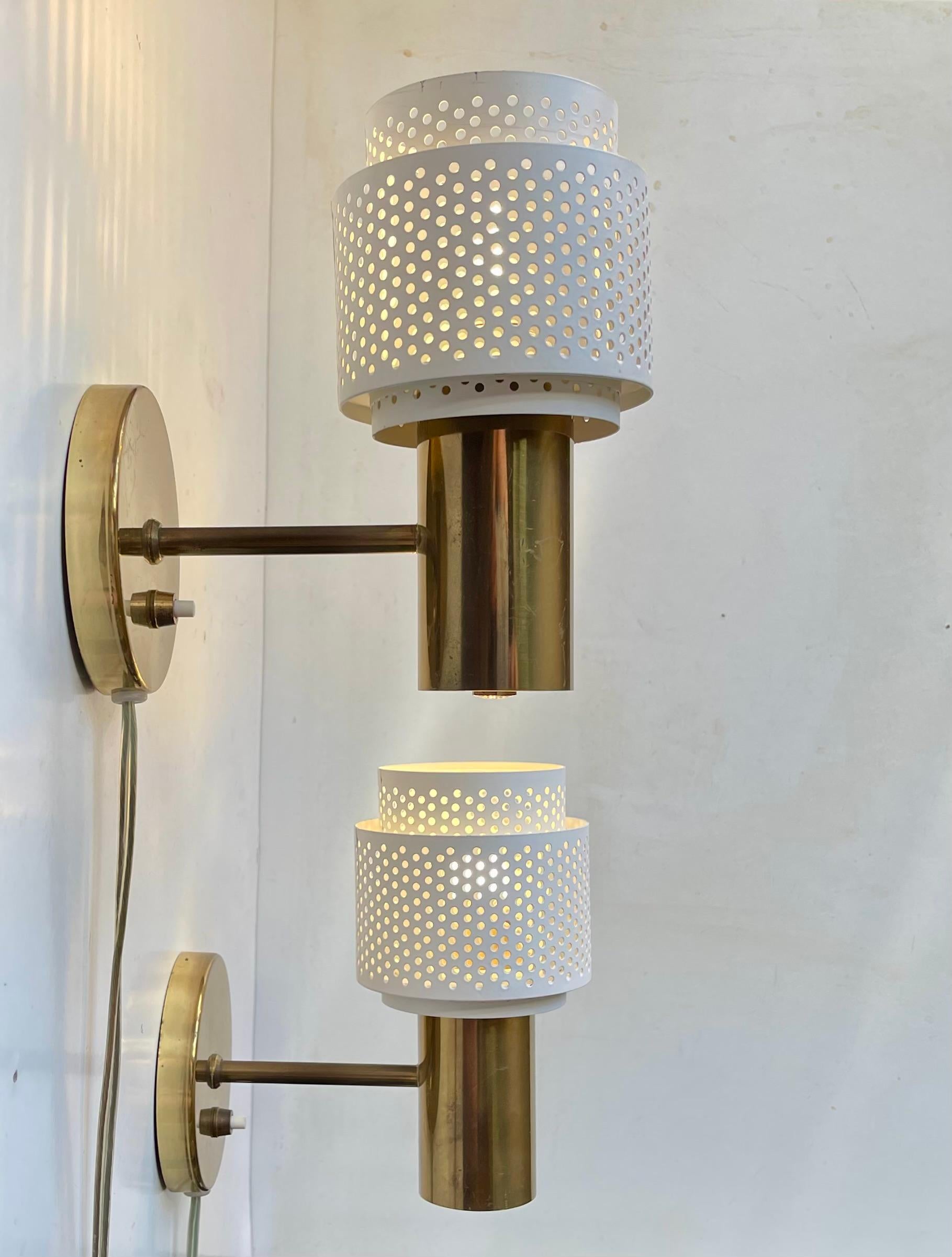 In the style of Hans Agne Jakobsson, Alvar Aalto and Pierre Guariche comes the stylish pair of up or down fixed wall sconces. They are fashioned from solid brass and features perforated white steel/iron shades. The set was made in Scandinavia during