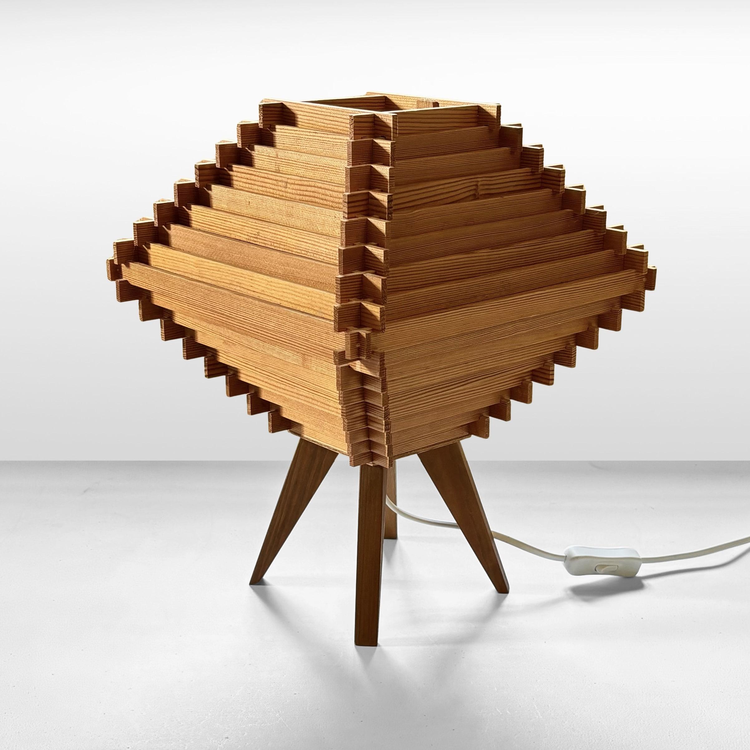 A rustic table lamp crafted from solid pine, featuring four triangular legs and a diamond-shaped shade constructed from wooden lamellas. Aesthetically in the manner of producers like Hans-Agne Jakobsson. A standard E27 lamp holder is placed at the