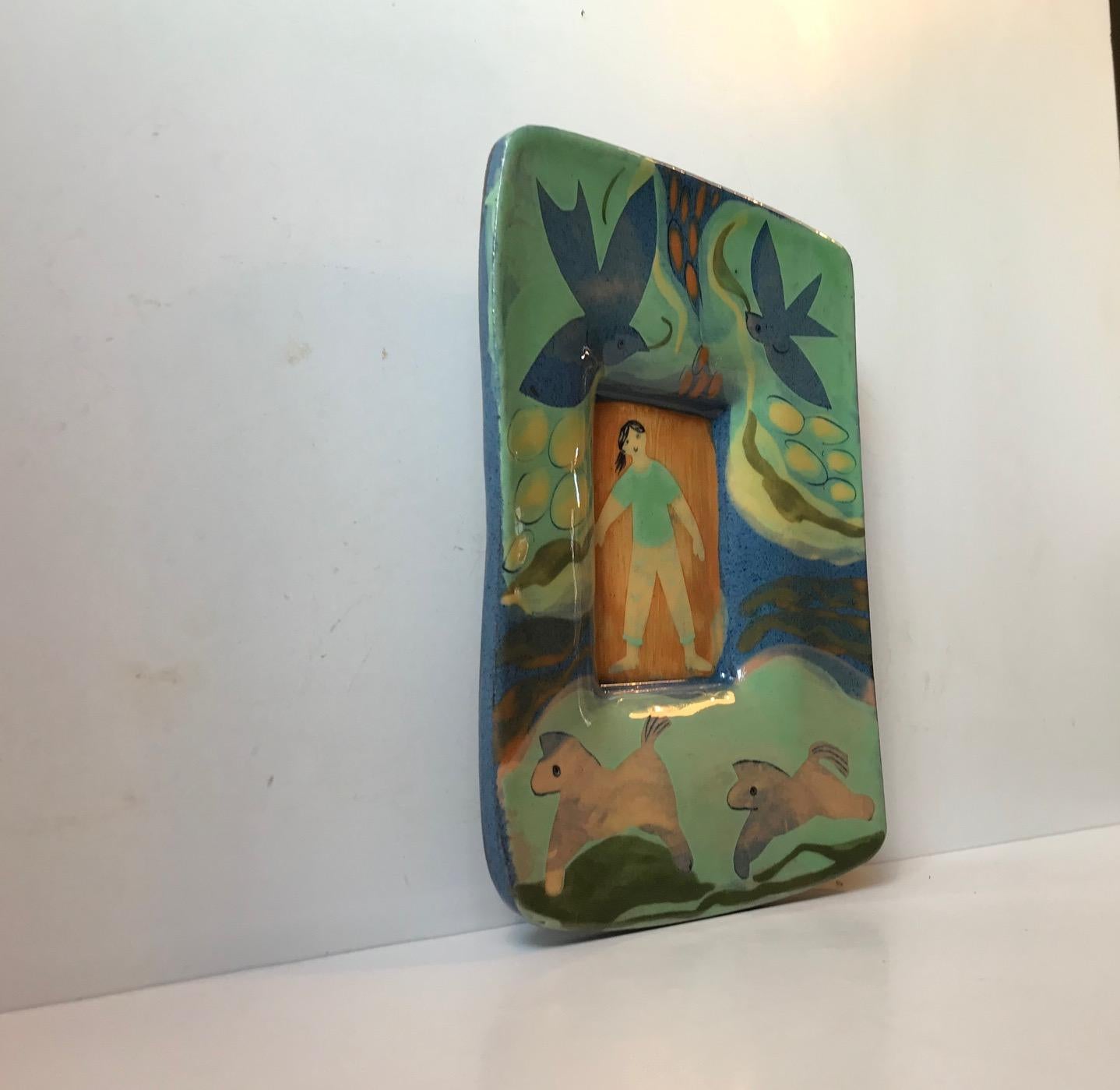 Vibrant and glossy in color palet this pottery wall plaque depicts a girl or woman lying in bed surrounded by naive dreamscape. It is executed in Scandinavia during the 1960s or 1970s by an anonymous artist or ceramist. It has an unidentified