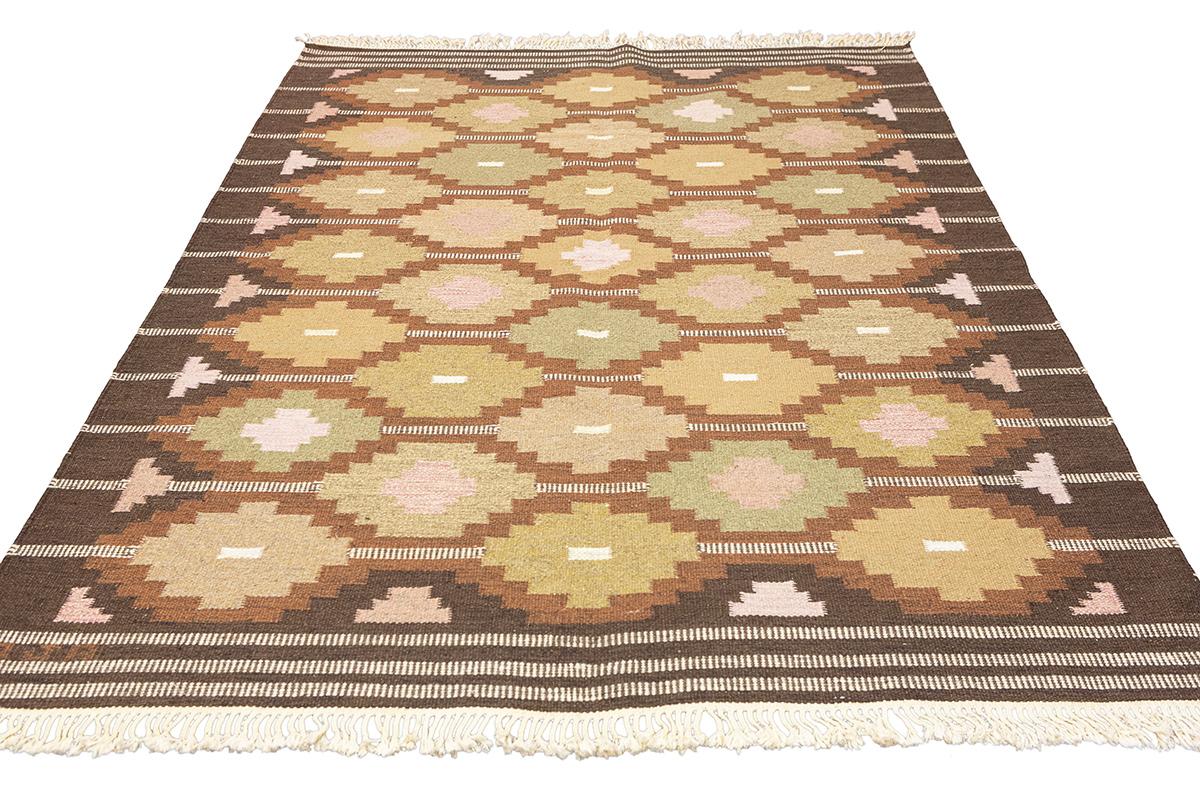 Questo tappeto scandinavo Rollakan è un ricco arazzo di tonalità terrose, che ricorda una foresta in autunno. Il suo motivo è un mosaico di toni naturali che porta in casa il calore e la serenità dell'aria aperta. Le forme geometriche tradizionali e