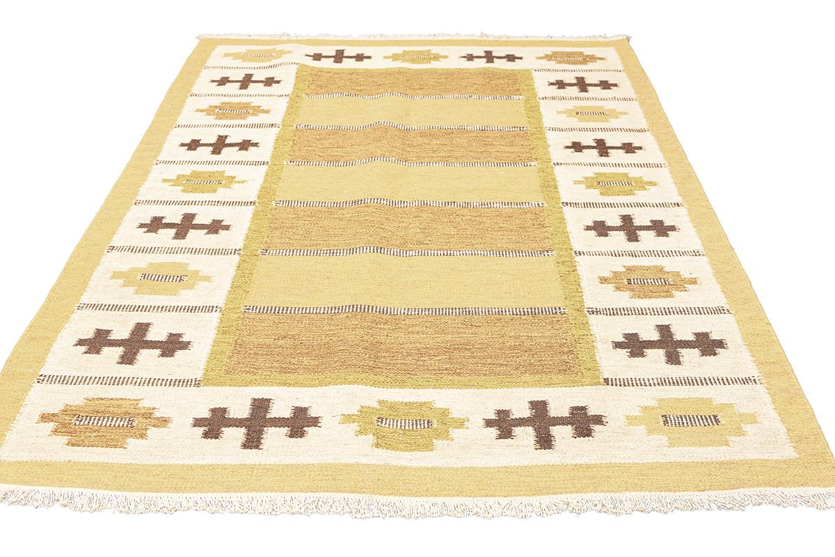 Questo tappeto scandinavo Rollakan è un vibrante tableau di toni terrosi, che intreccia il calore della terra illuminata dal sole e la sottile eleganza del design nordico tradizionale. Il campo centrale del tappeto è di un ricco giallo senape, che