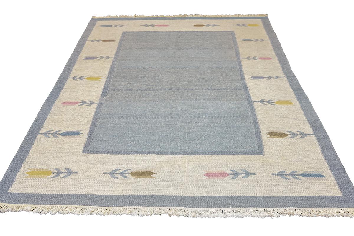 This Scandinavian Rug Rollakan is a stunning example of earthy elegance, woven with tones that bring to mind the tranquil beauty of a Nordic landscape at dawn. The central field of the rug is a soothing blue, reminiscent of a calm morning sky just