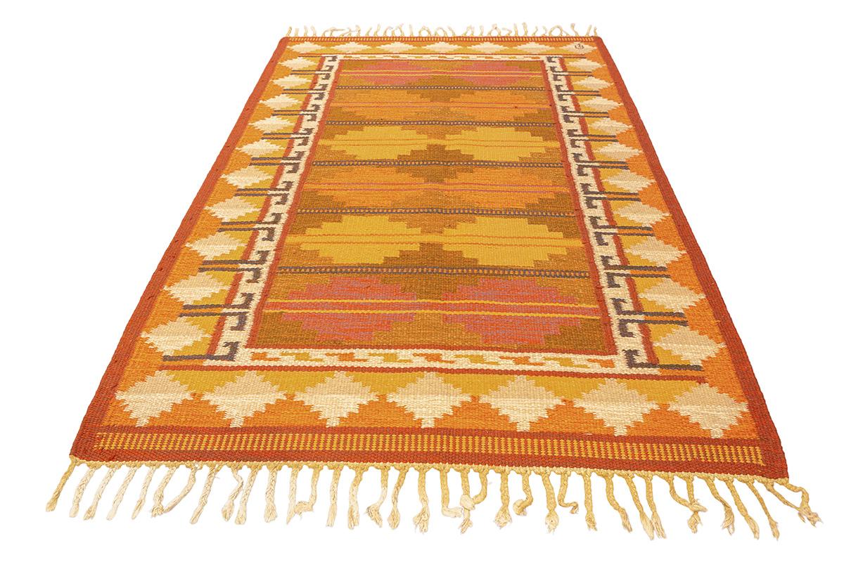 Questo straordinario tappeto scandinavo a trama piatta, che misura 201 × 139 cm, è una vivida celebrazione della tradizione del design nordico abbinata a toni caldi e terrosi. Tessuto con la tecnica del kilim, questo pezzo presenta una disposizione