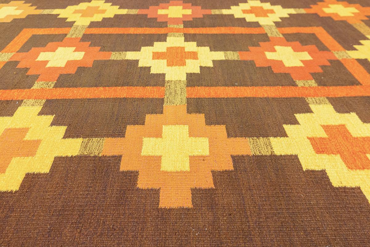 This Scandinavian Rollakan rug, measuring 167 x  243 cm, is a stunning example of Nordic craftsmanship with a bold geometric design and a warm earthy color palette. The intricate pattern consists of symmetrically arranged square motifs in shades of