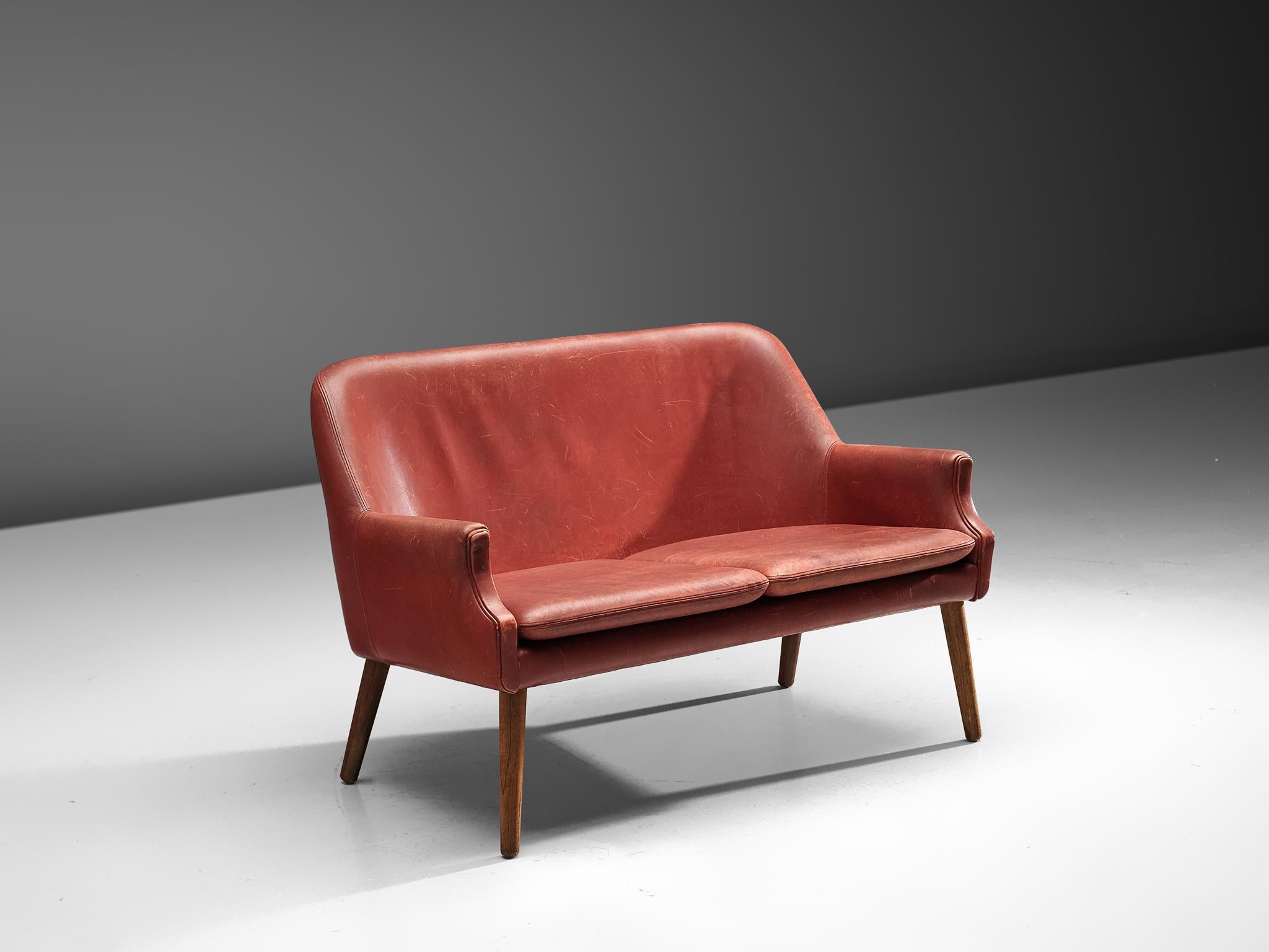 Scandinavian settee in red leather with wooden legs, 1950s.

Elegant two-seat settee in style of Danish designer Arne Vodder.
The high back is slightly curved end runs smoothly over into the slightly pointed armrests. The tapered wooden legs provide