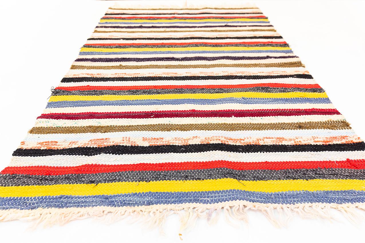 This Swedish kilim, a type of flatweave rug, features a striking linear design characterized by its horizontal stripes of varying widths and colors. The pattern is a harmonious blend of earthy tones like beige, brown, and cream, with bold accents of