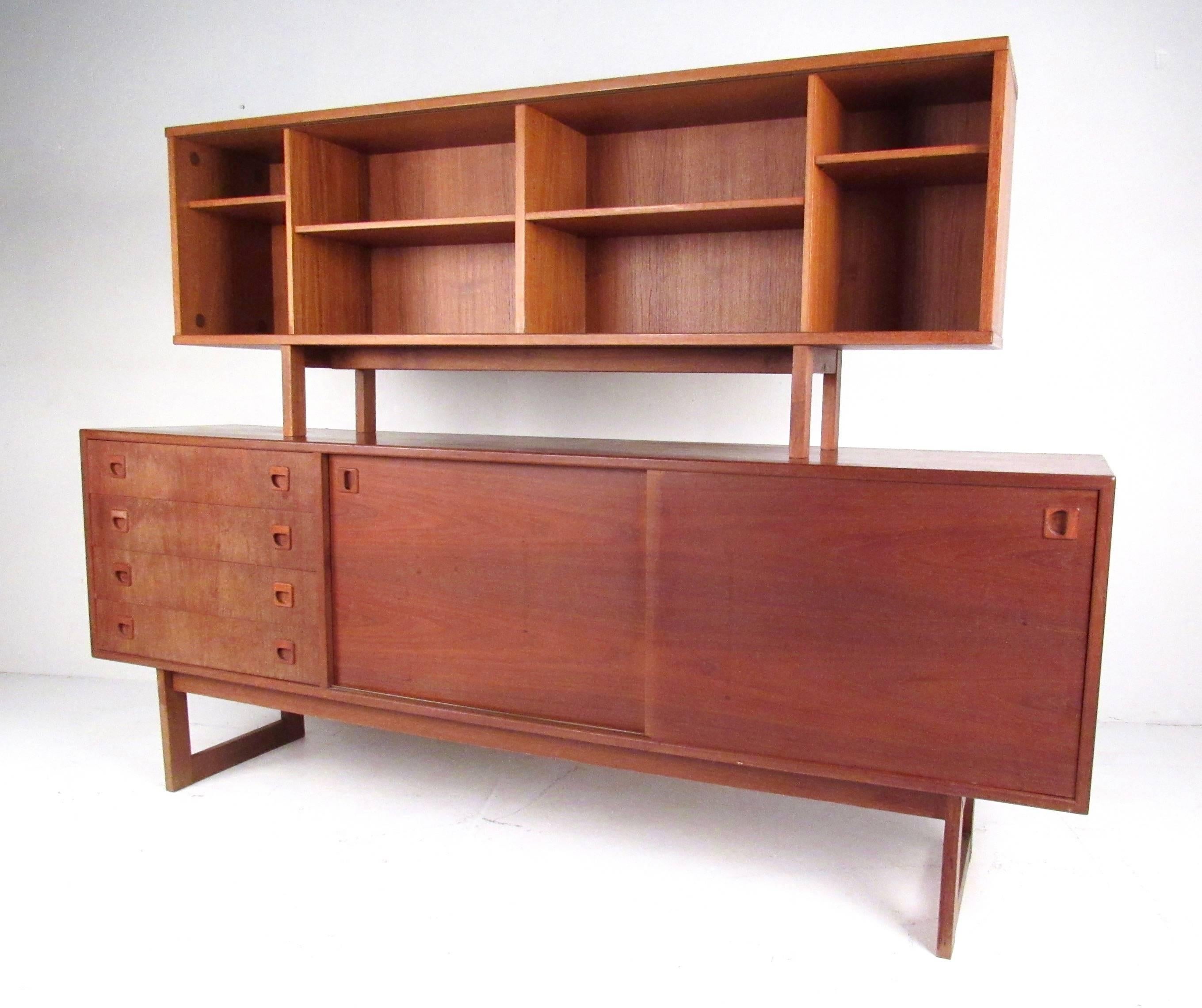Danish modern sideboard features vintage teak construction with dovetail drawers and sled legs. Spacious storage options include sliding door shelved cabinets, drawers with carved pulls, and bookcase display topper with glass doors. Please confirm