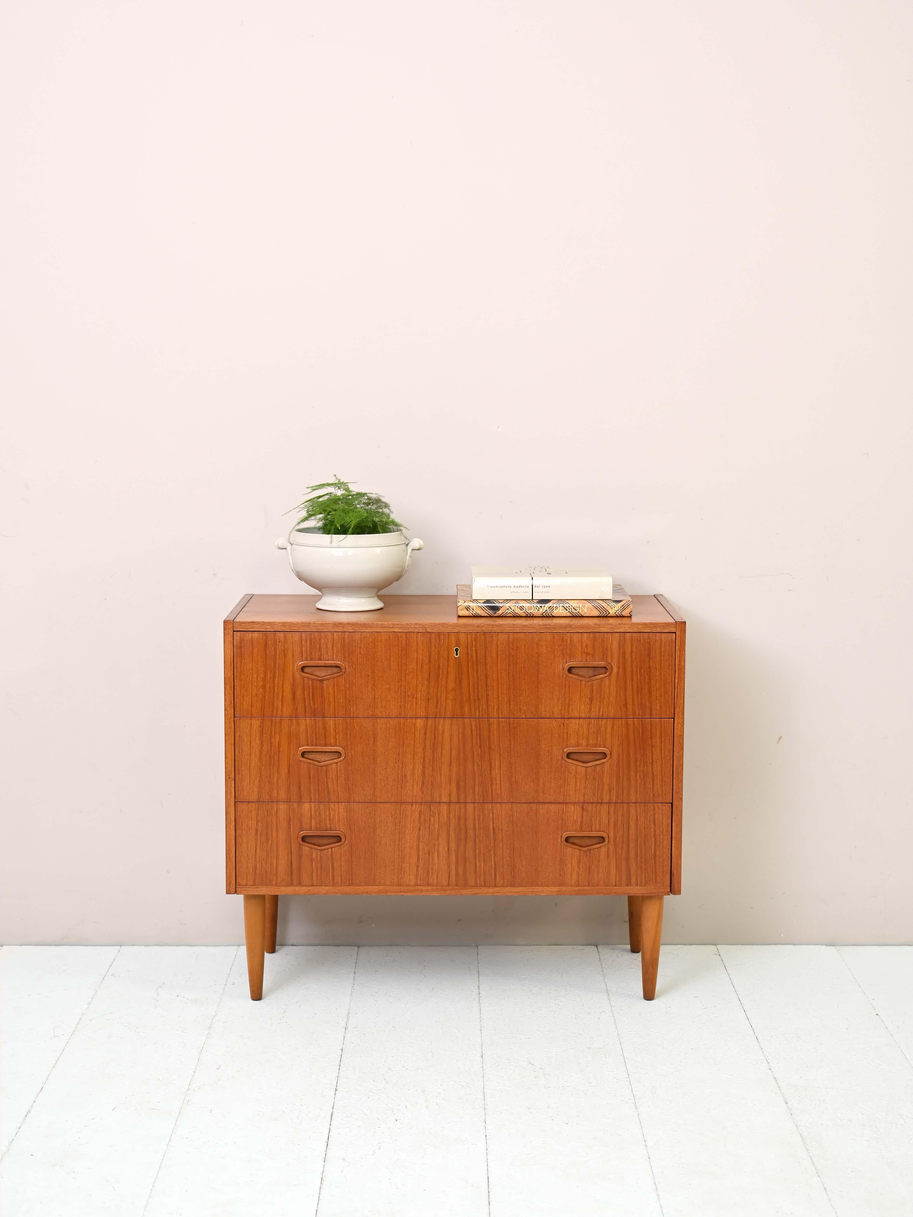 1960s teak cabinet with drawers.

A modern piece of furniture with a minimalist design. This chest of drawers consists of three drawers, the first of which has a lock. The handles are carved wood, in perfect Nordic style.
The tapered legs present