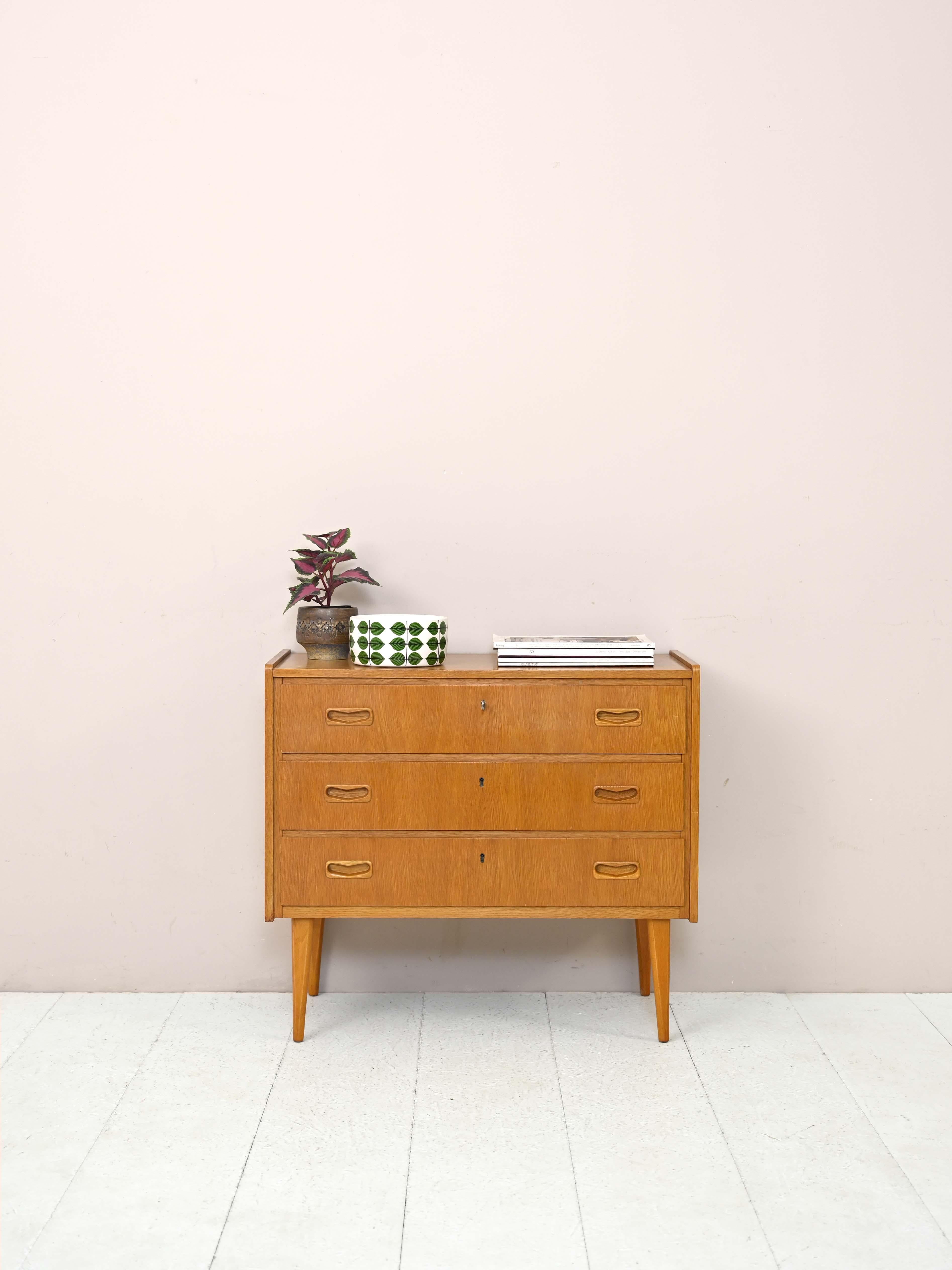 Modern 1960s teak cabinet.

A chest of drawers with modern and elegant lines that features refined details.
The drawers are lockable and feature a heart-shaped carved wooden handle.
Tapered legs slender the structure. Thanks to the golden color
