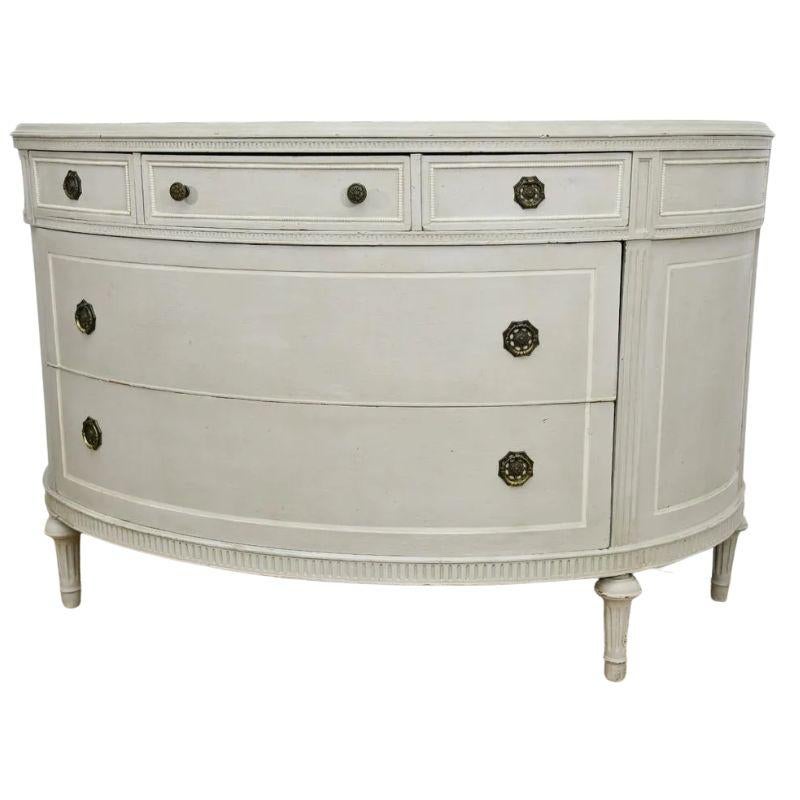 A beautiful white painted Swedish demi-lune cabinet with two large center drawers and three smaller drawers at top. The commode is fitted with octagonal hardware, straight fluted legs and carved detail to the borders and surrounding the drawers.