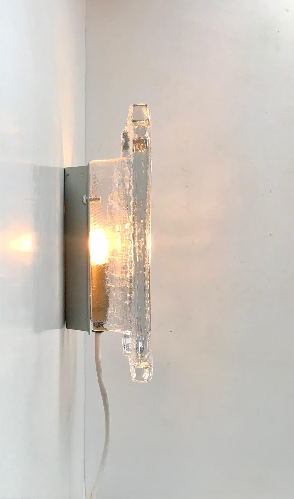 A post-Fagerlund wall light composed of thick ice-glass. Brilliant choice for hallway or cosy/ambient light. Manufactured and designed in-house at Orrefors in Sweden during the early 1970s. Measurements: 23.5 x 16 x 7.5 cm.