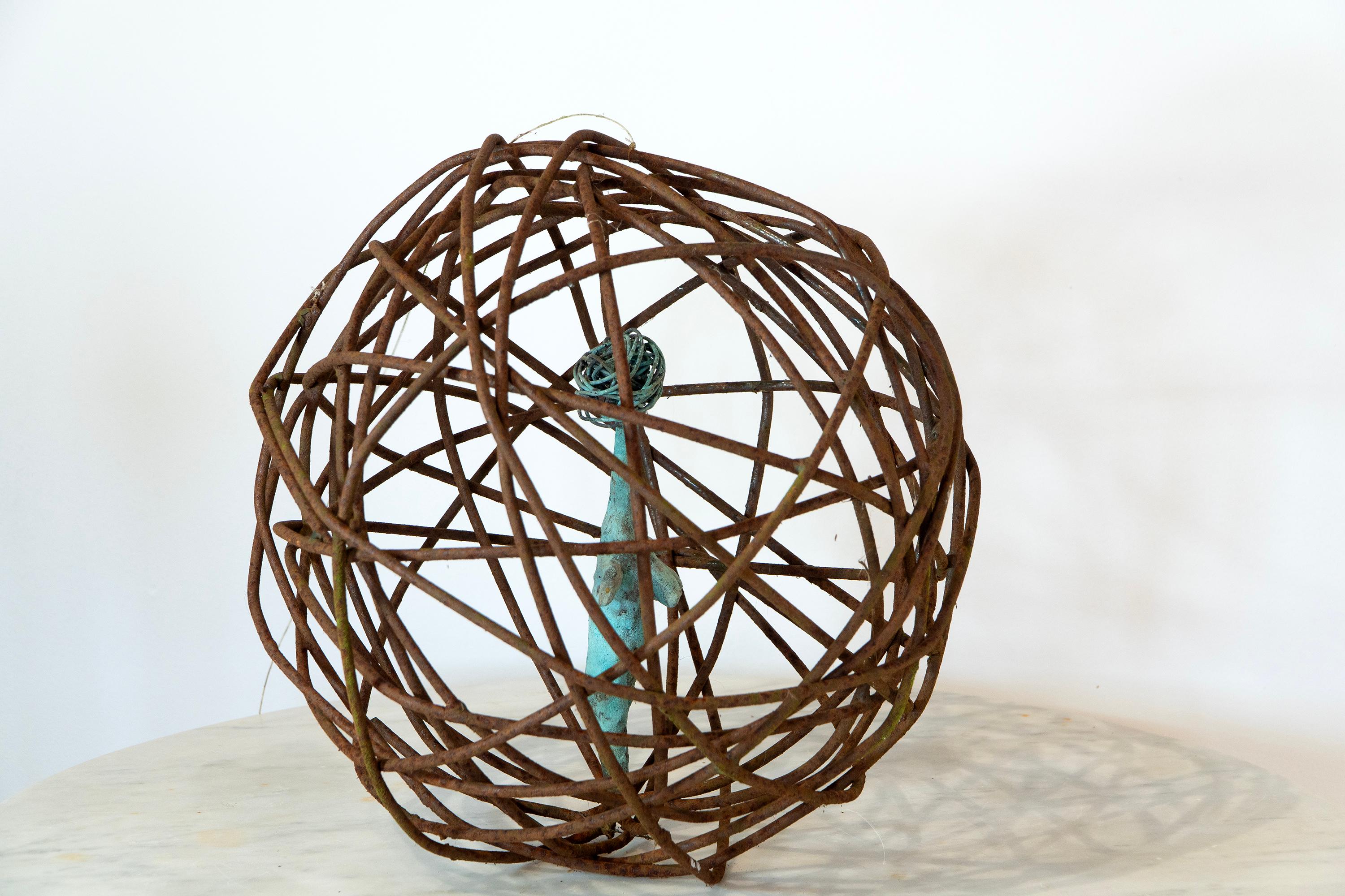 La figura in bronzo verdastro sospesa all'interno di una sfera scultorea in ferro arrugginito offre una meditazione poetica sul confinamento e sulla trascendenza. La Donna in Gabbia 2 (Ø 32 cm) di Aldonza è caratterizzata da un'esile forma ispirata