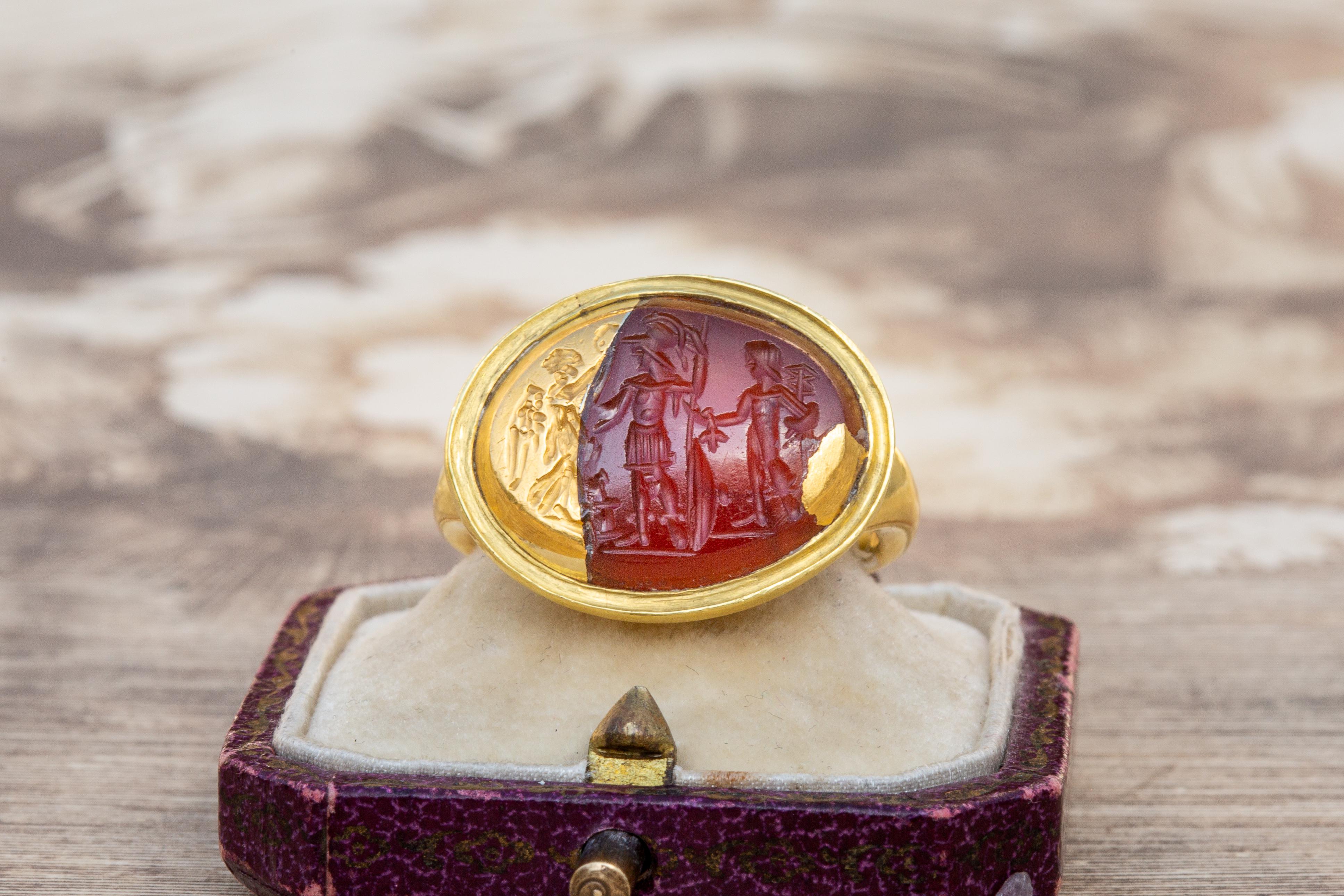 A superb, heavy high karat gold signet ring set with an ancient Roman intaglio, circa 1st century AD. The carved carnelian agate stone depicts two soldiers or warriors stood before a winged deity, most probably Nike, the Roman goddess of victory.