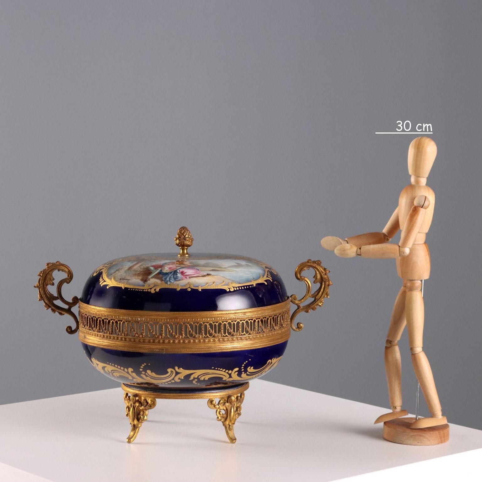 Scatola in porcellana e bronzo traforato con piedini e anse a volute fogliacee. Coperchio con riserve in oro a paesaggio lacustre e scena galante. Interno ornato con mazzetti di fiori. Sotto la base marchio della manifattura francese.