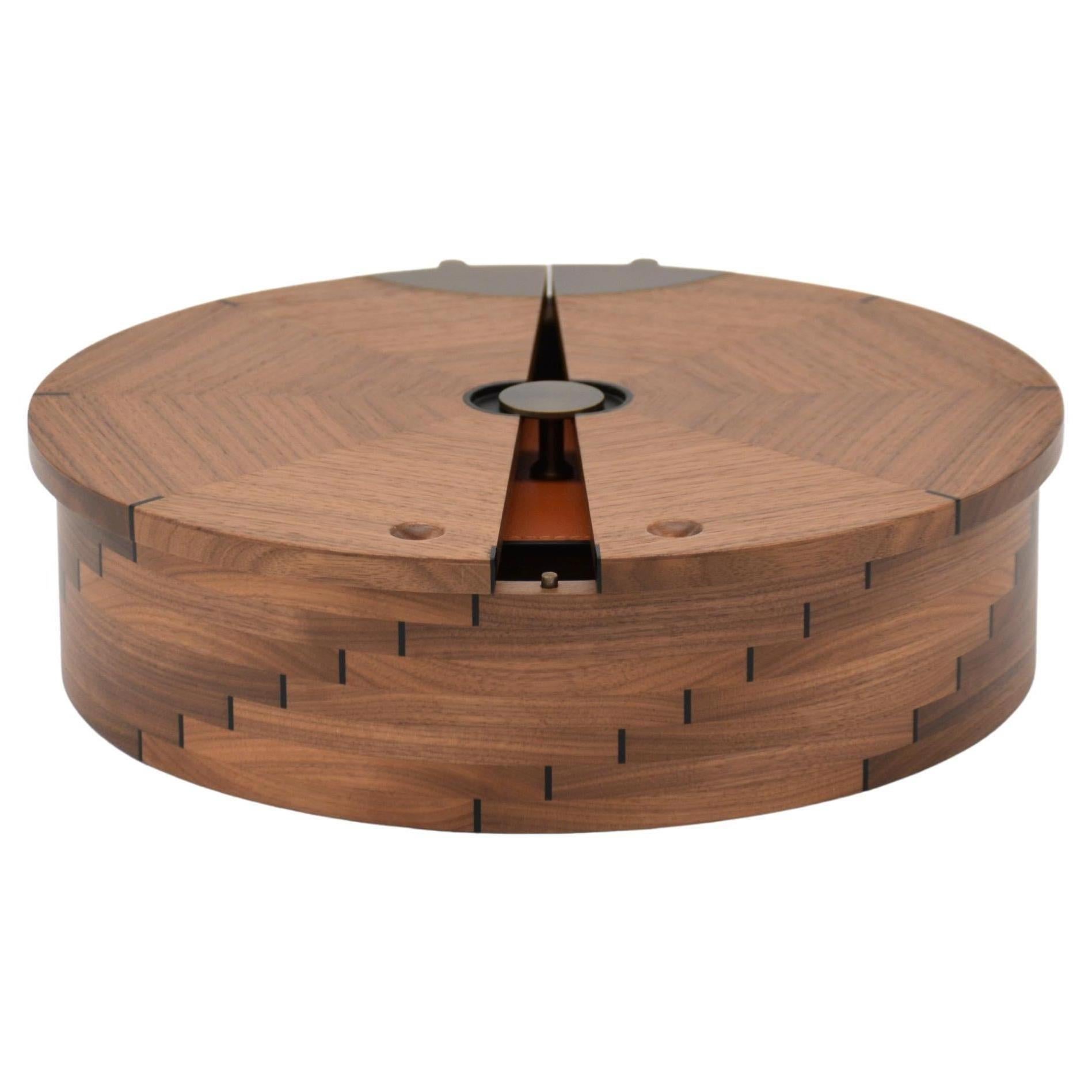 Round jewelry box made of solid turned walnut and decorated with small ebony inserts. The inlay forms an ascending geometric pattern. The lid opens by splitting into two parts by rotating on a bronzed brass pin and simulating the movement of a