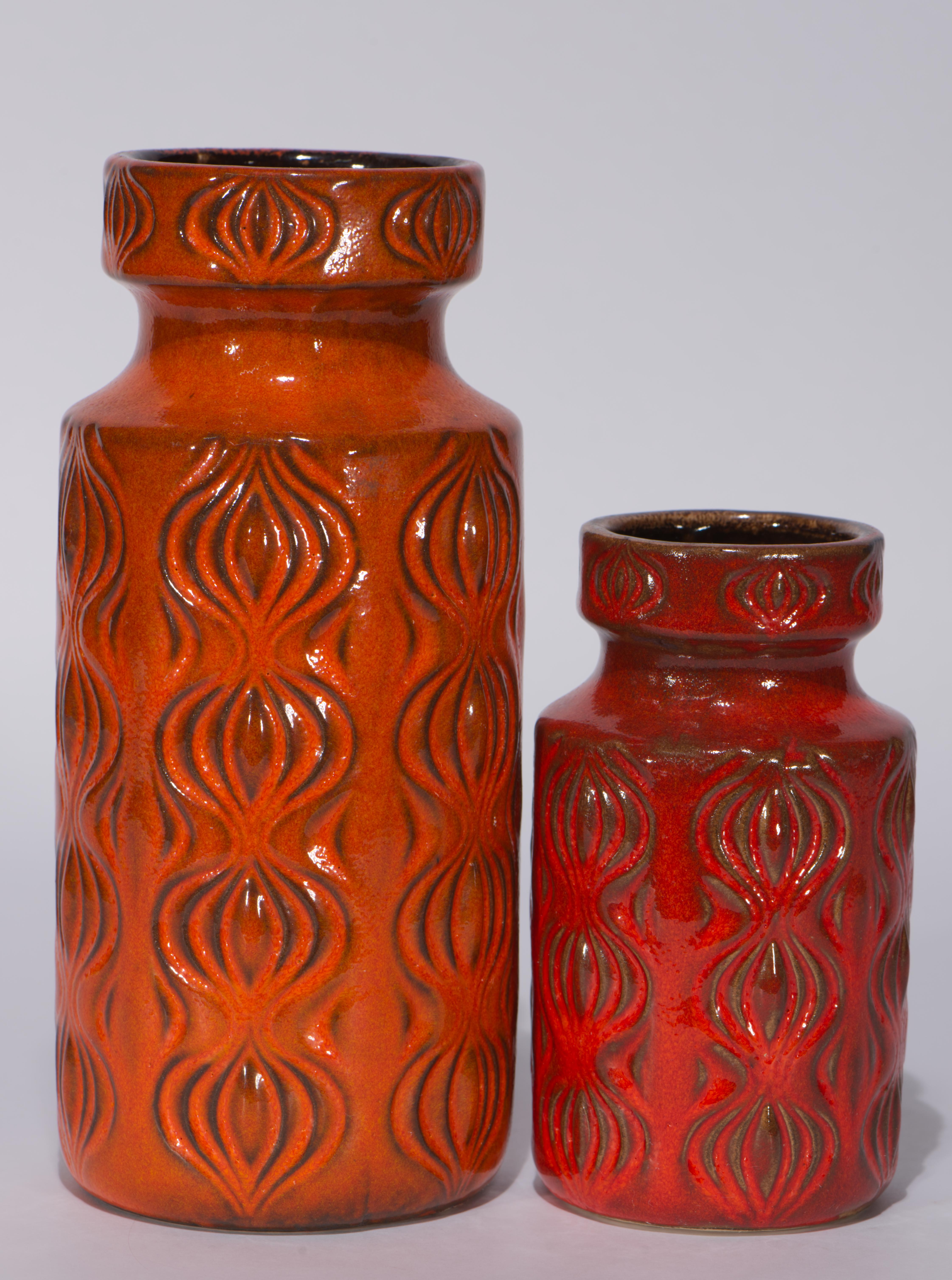 El juego de 2 jarrones vintage de West German Pottery presenta decoración moldeada de Ámsterdam, a menudo llamada Cebolla, y esmalte semimate de textura de Lava Grasa en paleta de rojos y naranjas. Ámsterdam, presentada en 1972, se caracteriza por