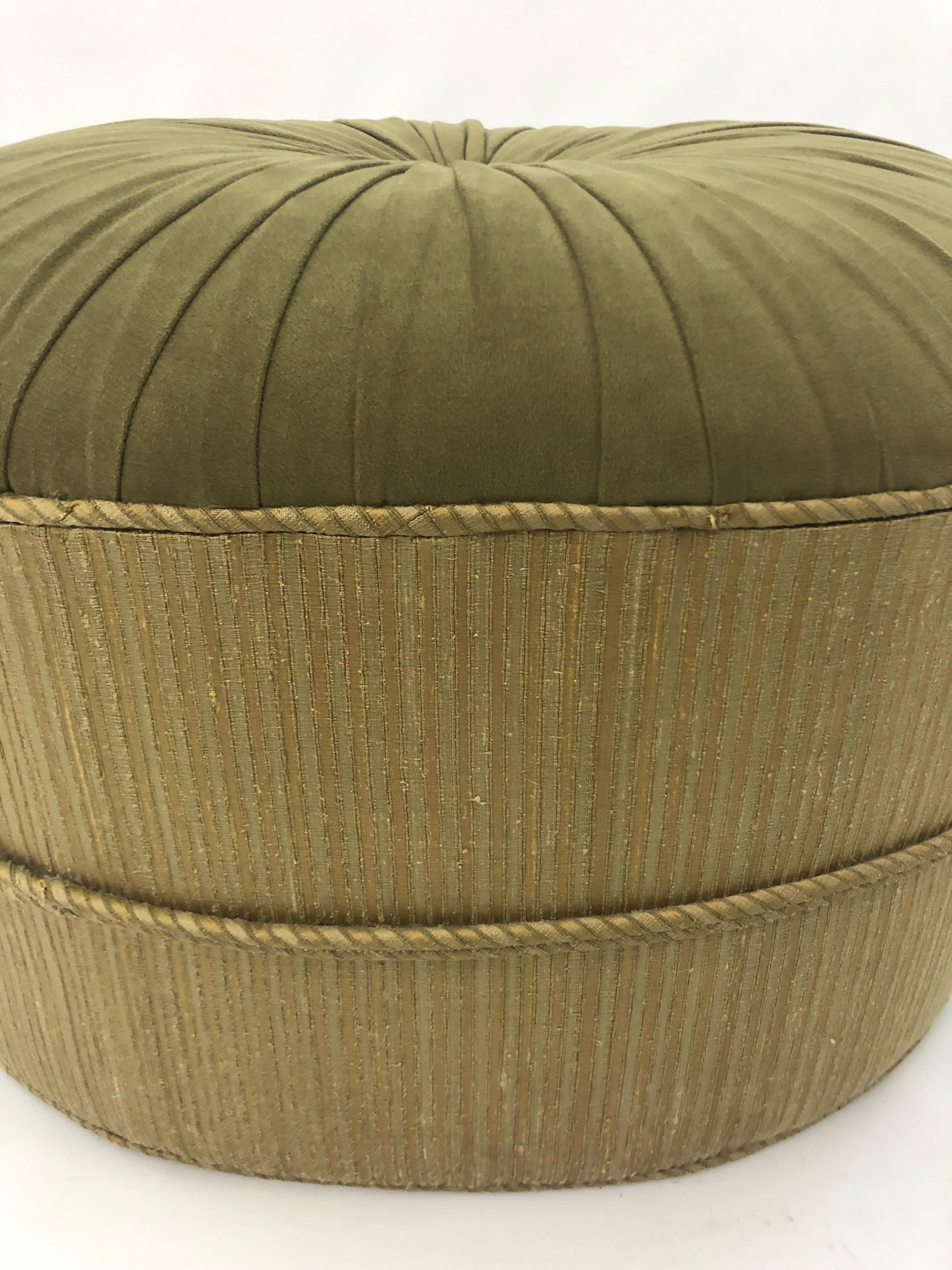 A stylish oval pouf or ottoman in a wonderful olive green ultrasuede having single button and pleating on top and a lighter color of greenish taupe around the periphery.