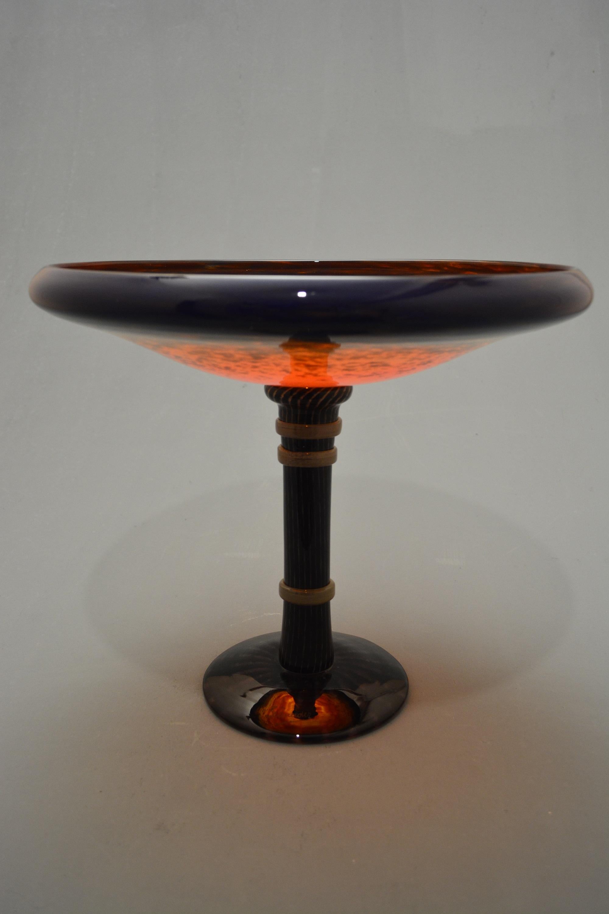 


Bowl with curving rim in blue powdered glass, changing to red- orange in the center; stem in violet powdered glass with yellow stripes, encircled by three yellow applied rings; violet foot with yellow stripes.
Schneider production.
Signed