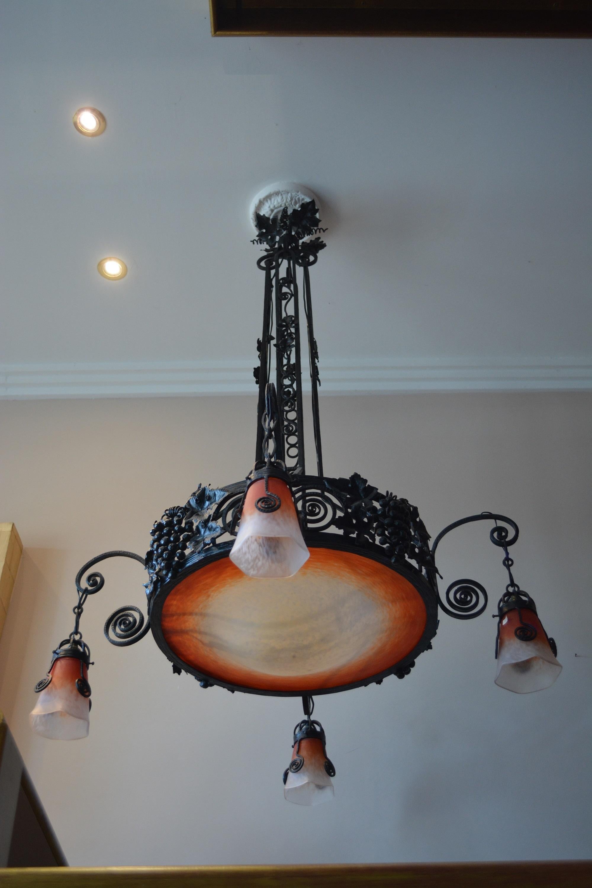 Five lights chandelier with a very large central cup in orange-white powdered glass.
The wrought, iron setting is decorated white grapevine leaves, fruits, and spirals.
Four arms with spirals motif as a support of four tulips.
The wrought, iron