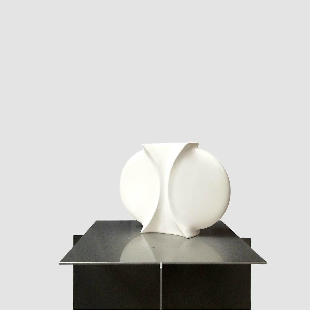 Le *Vase Schol* (1987) de Jan van der Vaart, réalisé en porcelaine émaillée blanche, illustre la maîtrise de l'artiste en matière de formes minimalistes et géométriques. Mesurant 24 x 18 x 7 cm, ses surfaces lisses et incurvées et son élégante