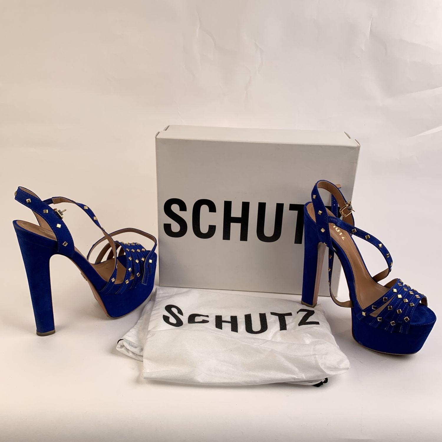 Zapatos de tacón Schutz de ante azul. Llevan adornos de tachuelas metálicas doradas, diseño de plataforma y correa ajustable en el tobillo. Plataformas de 5,6 cm. Tacones de bloque cubiertos (15,8 cm). Talla: EU 39, BRA 37, US 8B (La talla mostrada