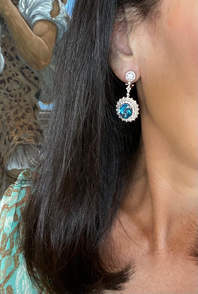 Exquisite pair of estate, oval shaped faceted blue topaz sparkling gemstones are talon prong set in 18 karat white gold and surrounded by 5.26 total carats of round brilliant cut diamonds. Earrings are for pierced ears and have posts and omega lever