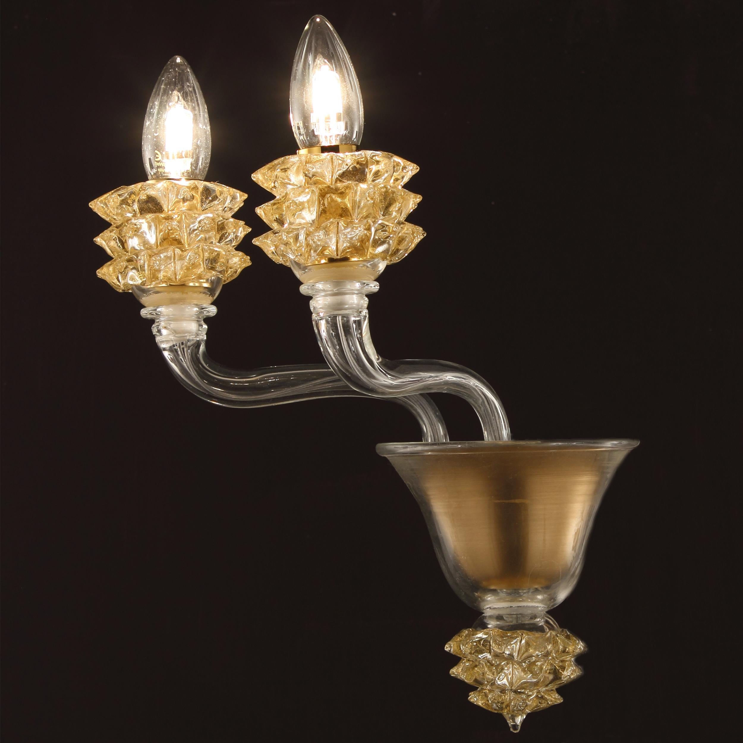 L'applique Diamante avec 2 bras en verre Murano transparent et détails dorés est un produit bien proportionné.
Le verre des bras est lisse. Les éléments remarquables de cette applique sont les coupes, qui sont créées selon un processus de